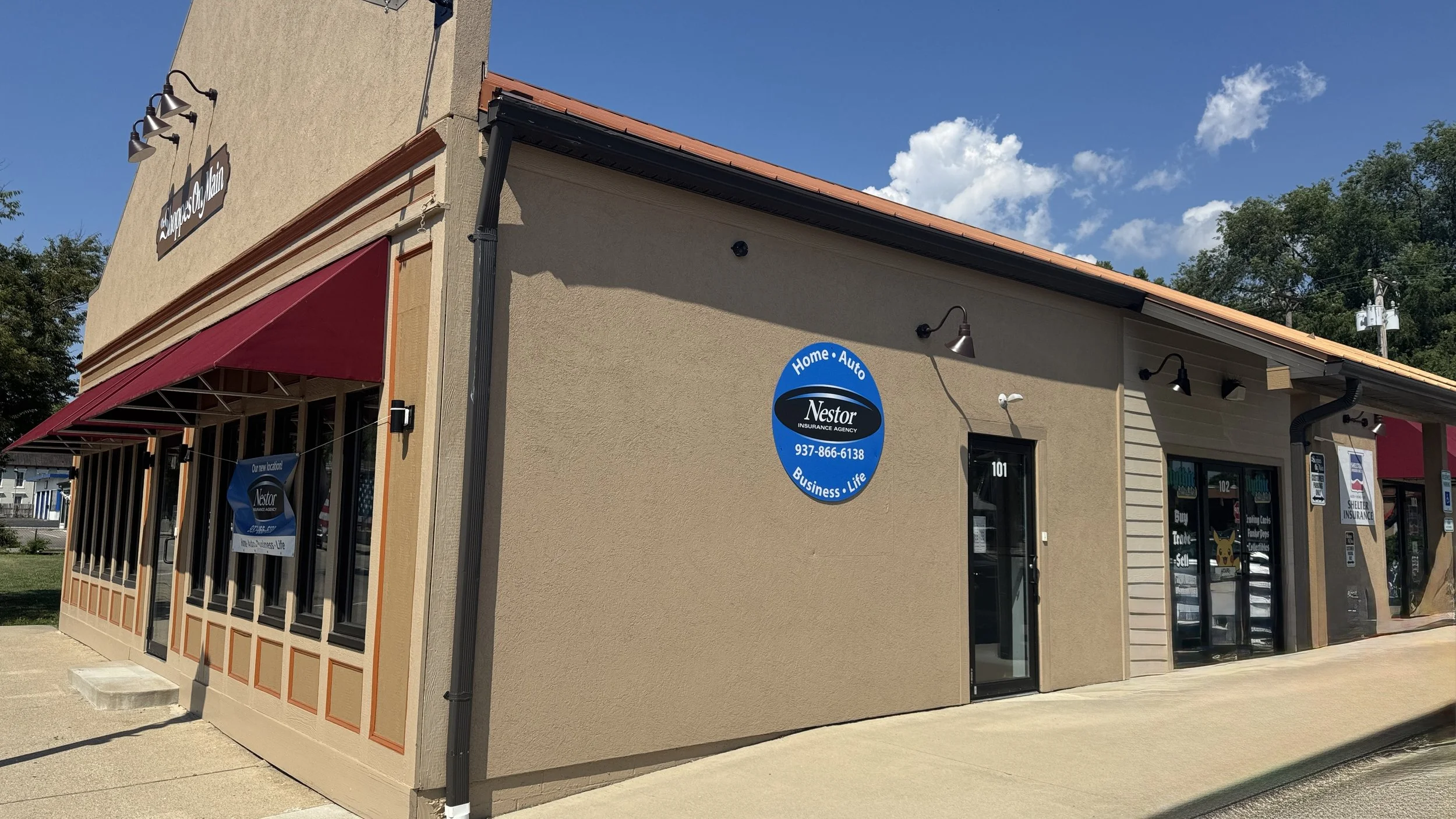 Exterior view of a beige commercial building with red awnings and multiple windows, a blue sign for Nestor Insurance Agency, and a street number 101. Clear blue sky with some clouds in the background.