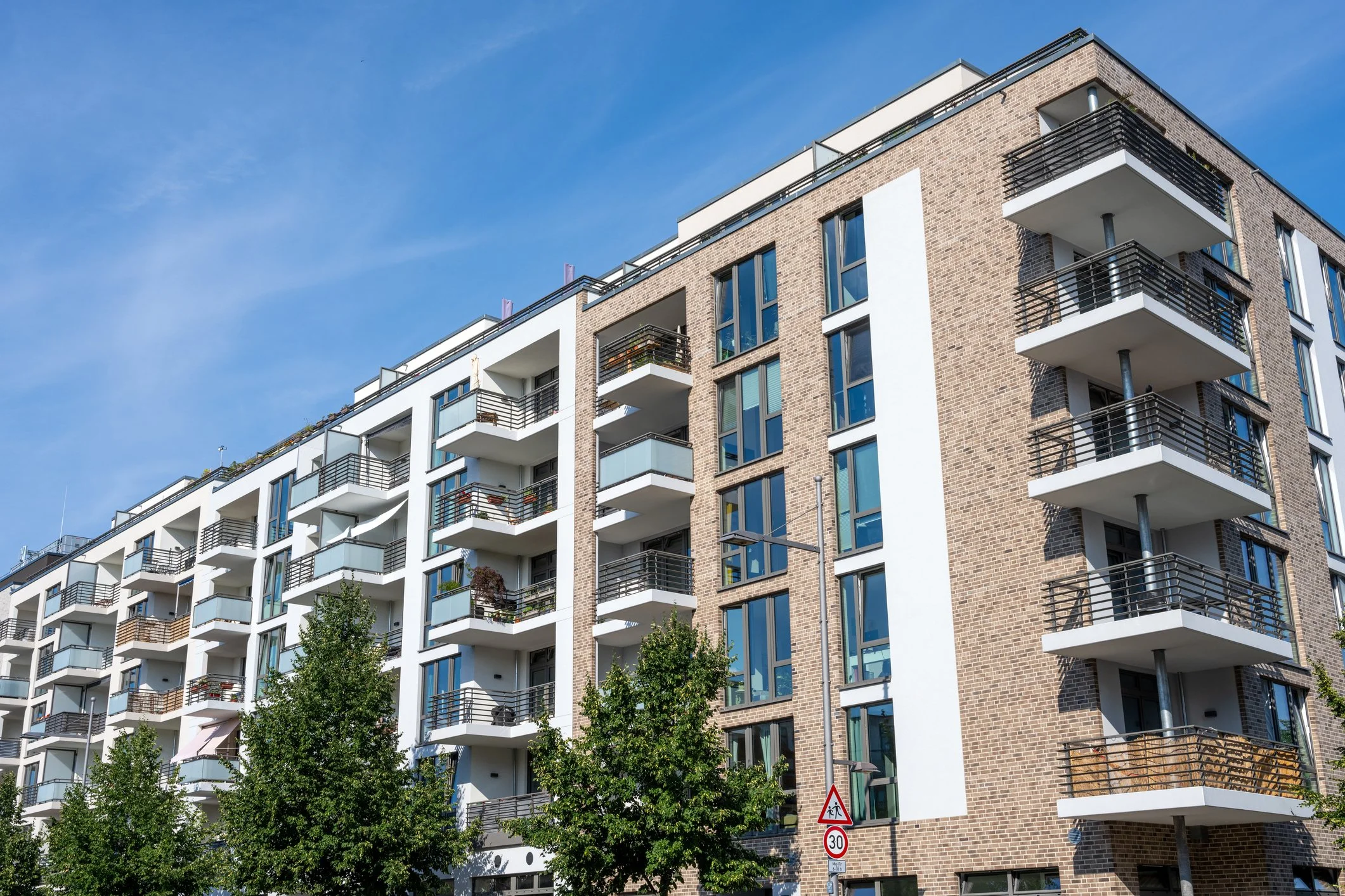 Modern multi-story apartment building with brick and white exterior, multiple balconies with black railings, and large windows, under a blue sky.