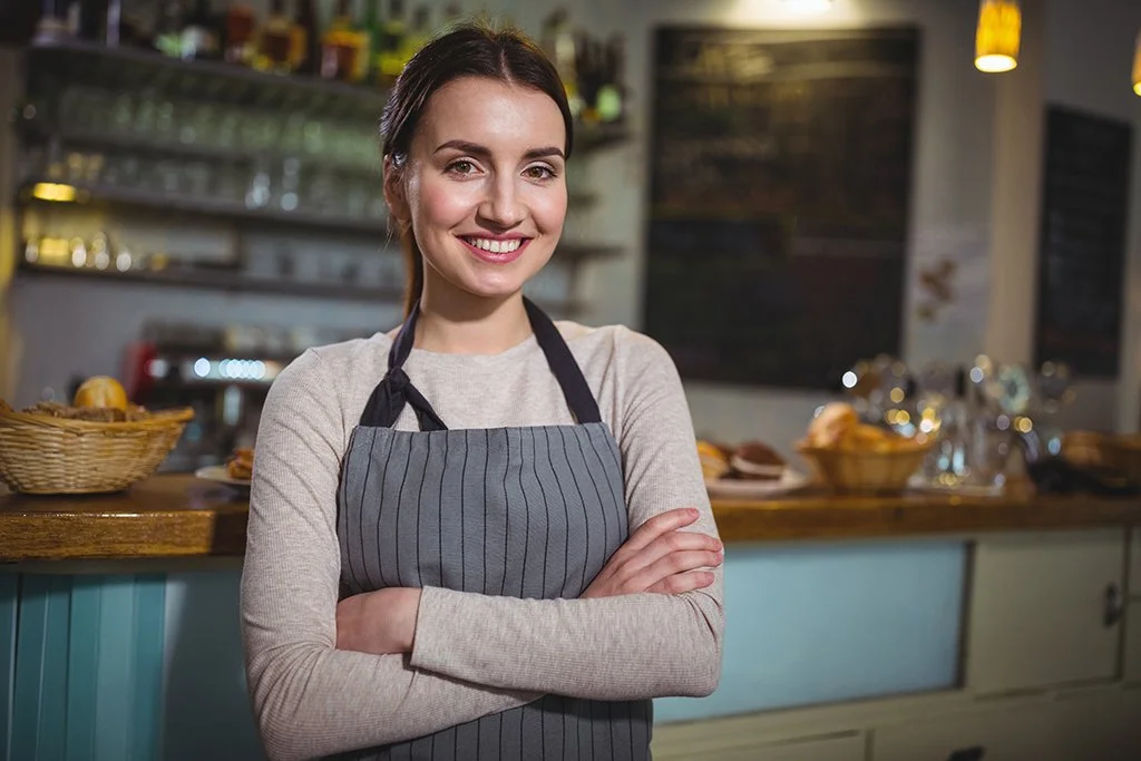 Smiling woman in a gray apron standing with arms crossed behind the counter of a cafe or bakery, with baked goods and a chalkboard menu in the background.