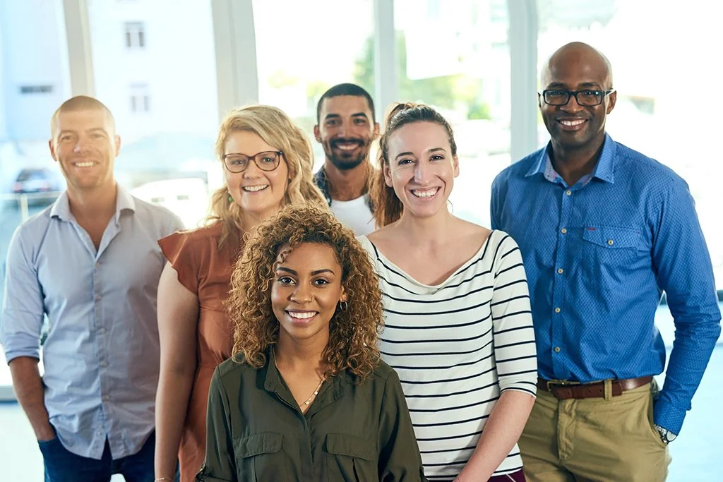 Group of six diverse smiling people standing in a bright office with large windows