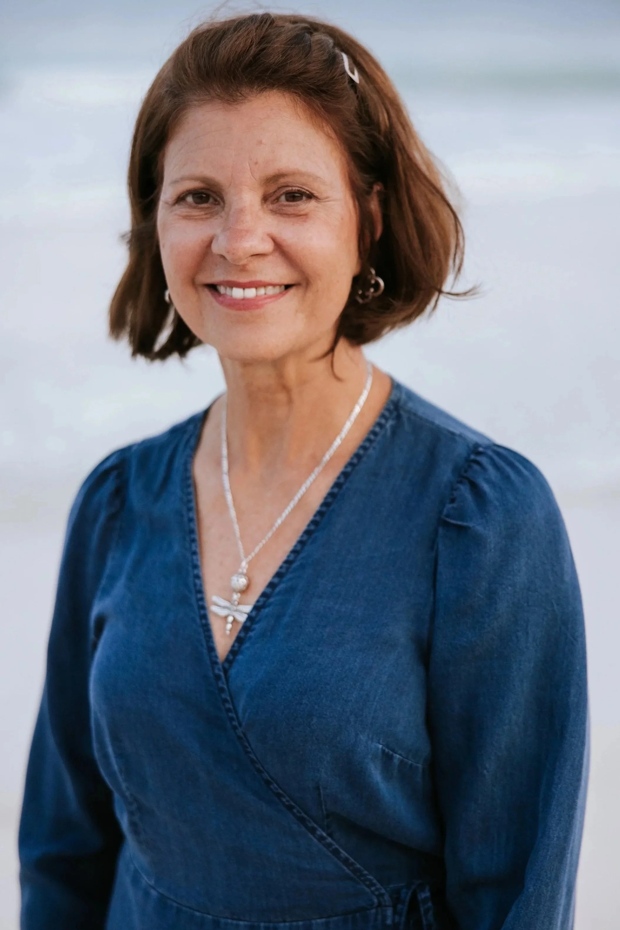 A woman with short brown hair, wearing a denim top and silver jewelry, smiling against a blurred outdoor background.