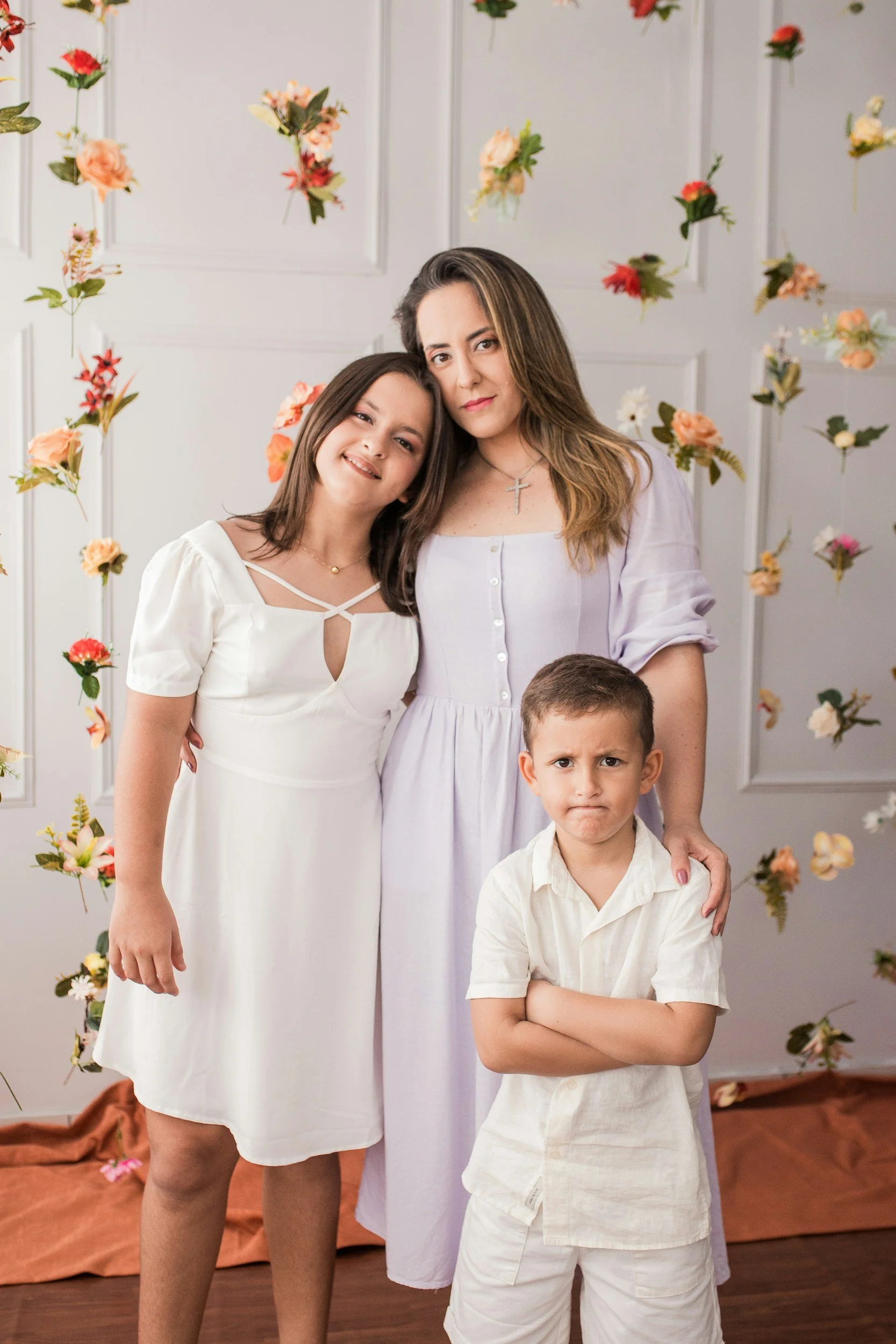 A woman with long brown hair wearing a white dress stands next to a girl with shoulder-length brown hair in a white dress with crisscross straps, both smiling and leaning their heads together. A boy with short brown hair wearing a white shirt and shorts stands in front with a serious expression and crossed arms. The background features a white wall with panels and hanging floral decorations.