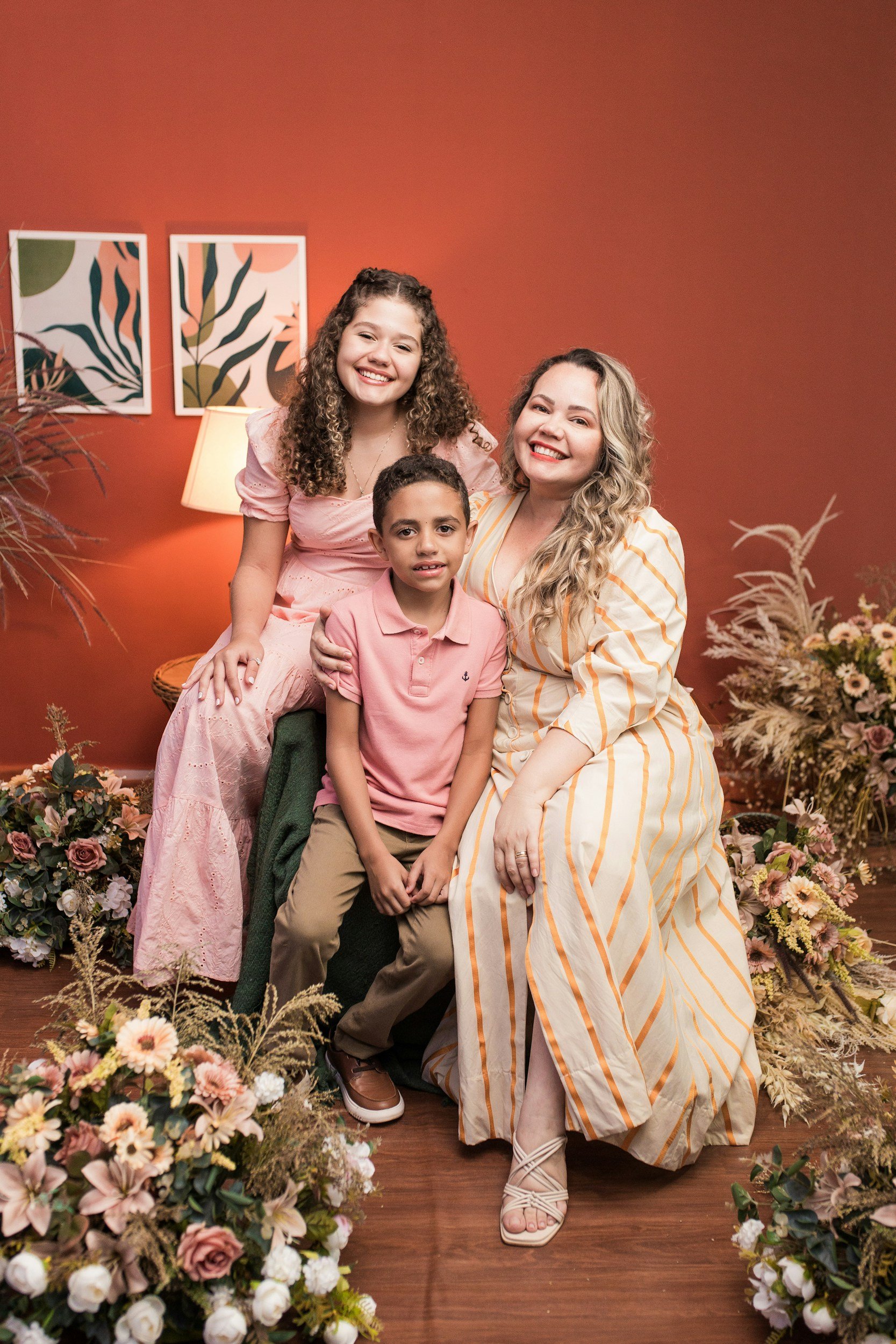 Family of three posing indoors with a floral arrangement and a red wall background, smiling at the camera. The woman has blonde curly hair, the girl has long curly hair, and the boy has short dark hair. They are dressed in pastel and light-colored clothing.