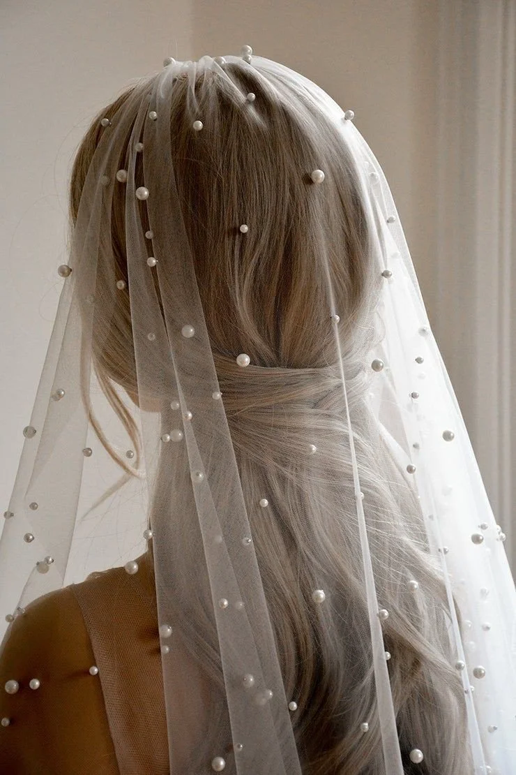 A bride with long, wavy blonde hair wearing a lace veil decorated with pearls.