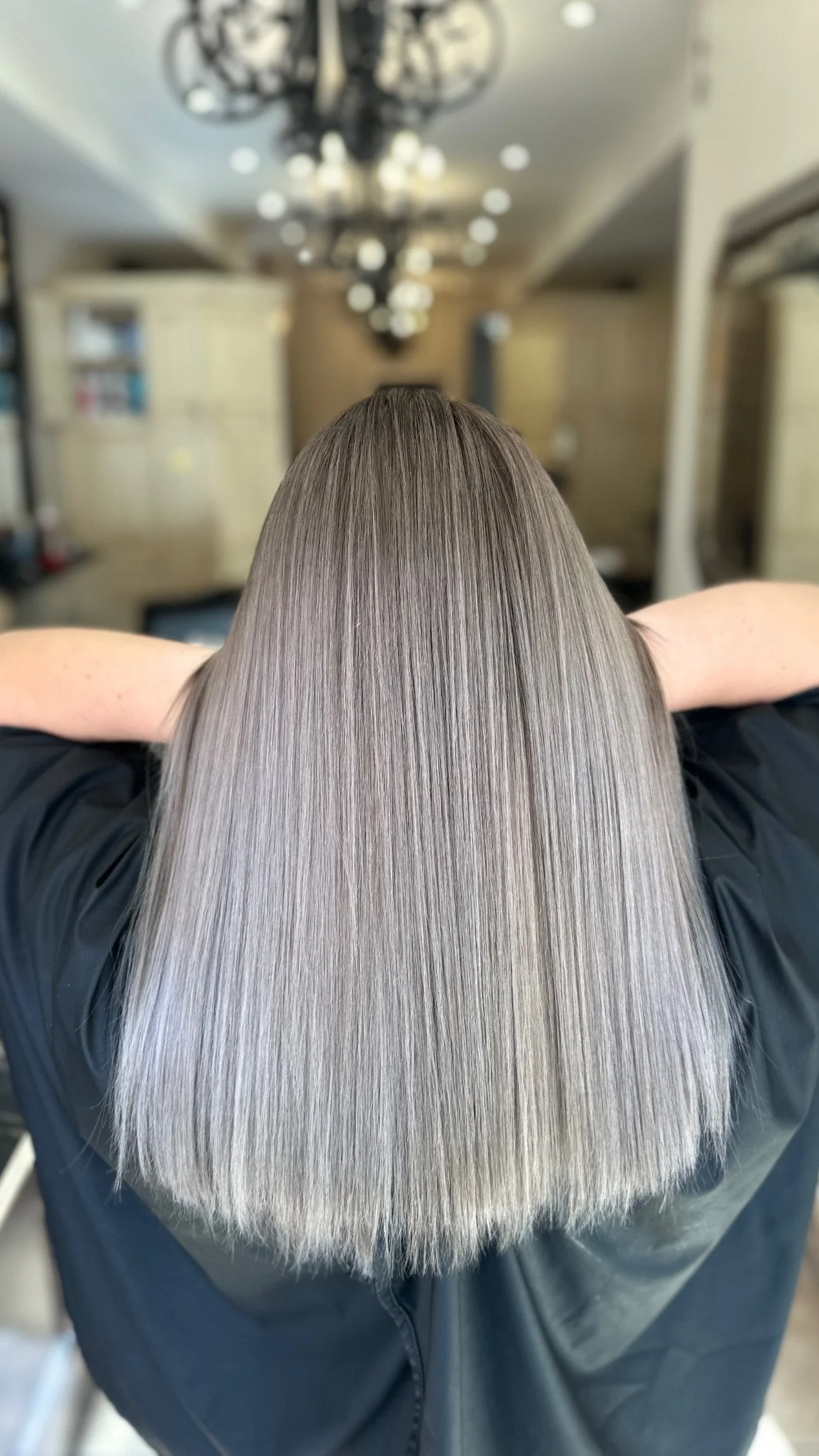 Person with long, straight, light gray hair or wig, wearing a black outfit, in a salon with chandeliers and chairs.