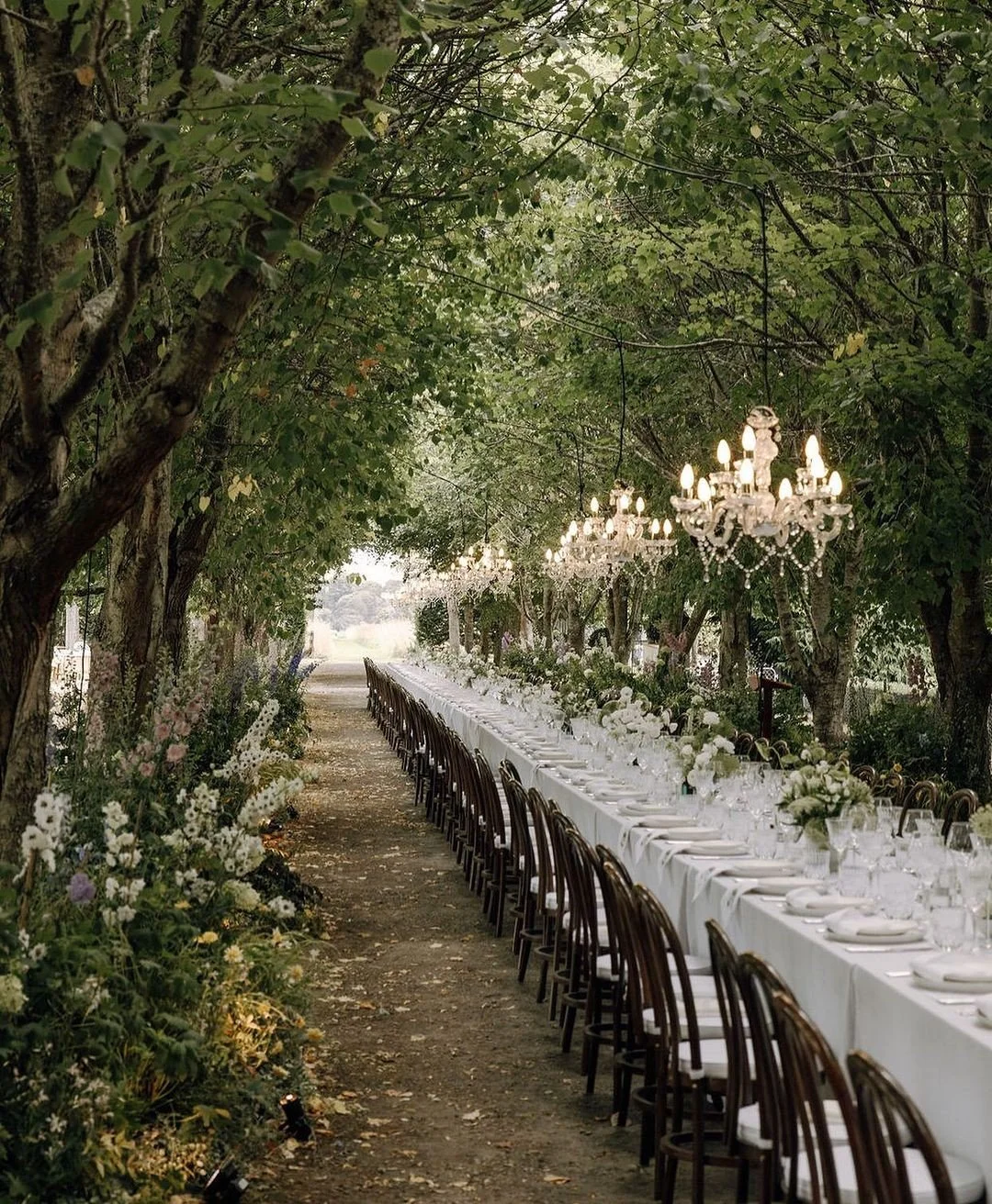 An outdoor banquet setup under a canopy of trees with a long white table, white tablecloth, elegant chandeliers hanging from branches, and floral arrangements, creating a tranquil and sophisticated atmosphere.