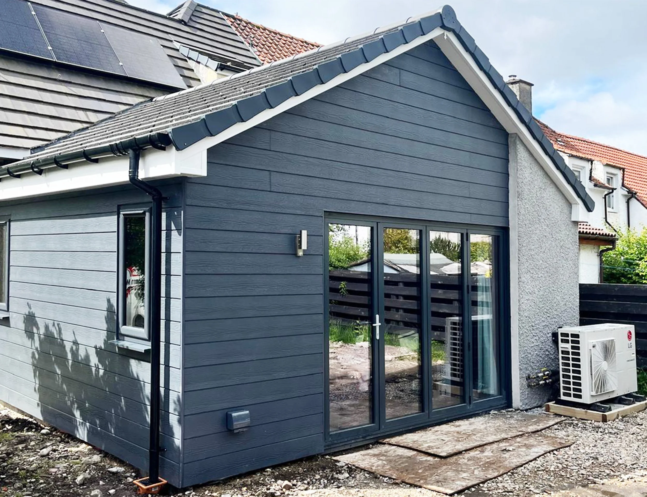 A small modern house or extension with blue exterior siding, a gabled roof with black shingles, large glass sliding doors, and an outdoor air conditioning unit, situated on a gravel yard.