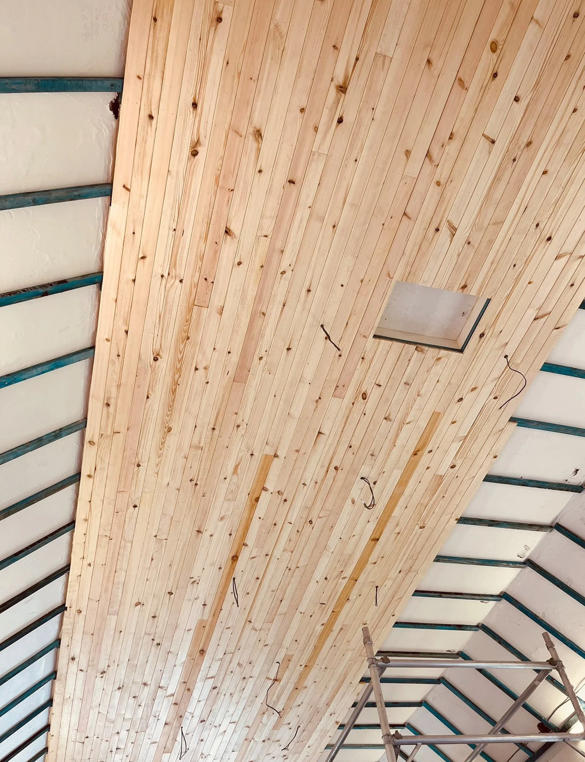 Interior view of a ceiling under construction with wooden planks, a square cutout, metal framing, and a scaffold.