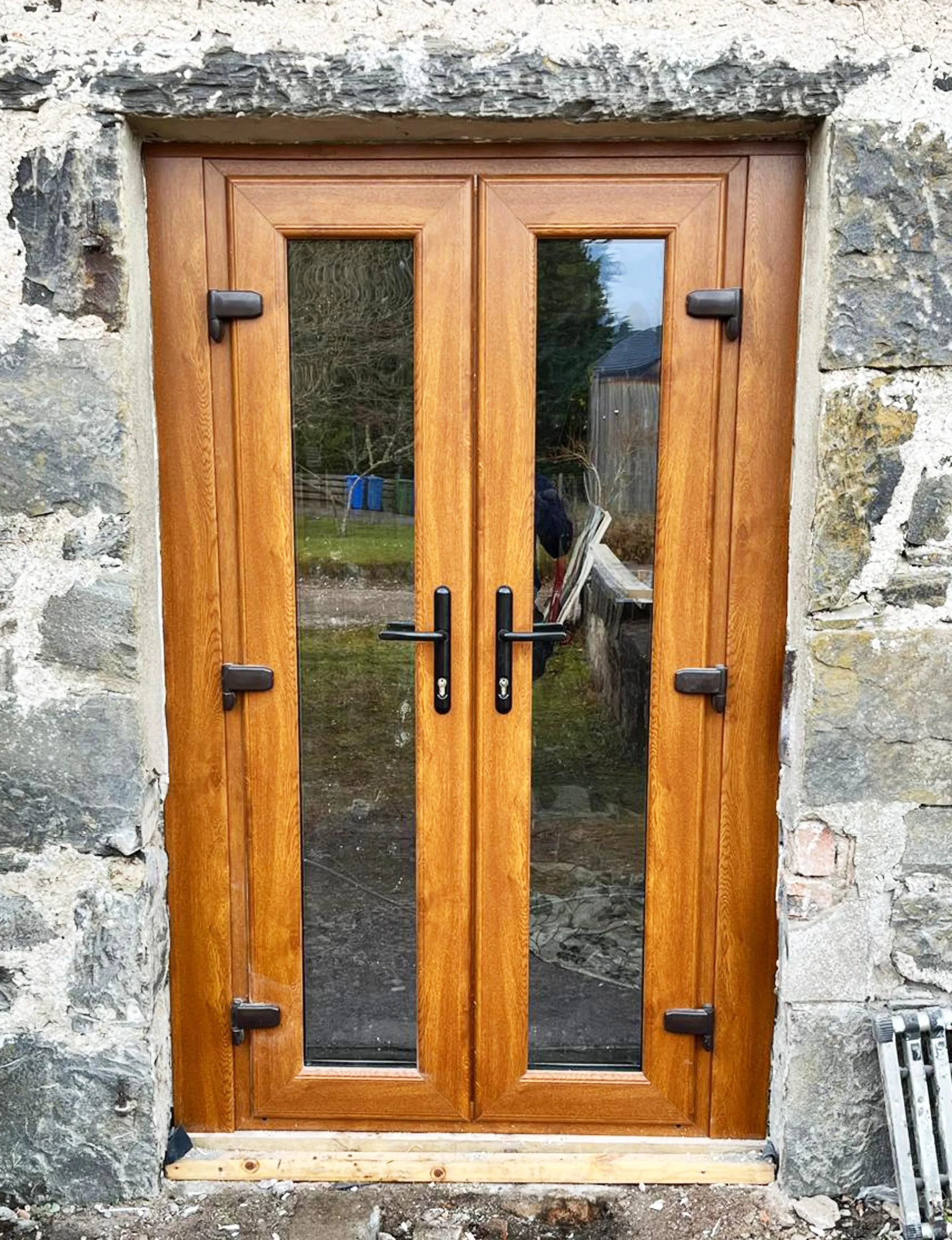 Wooden double door with glass panels, black handles, and locking mechanisms, set in a stone wall with visible construction details at the bottom.
