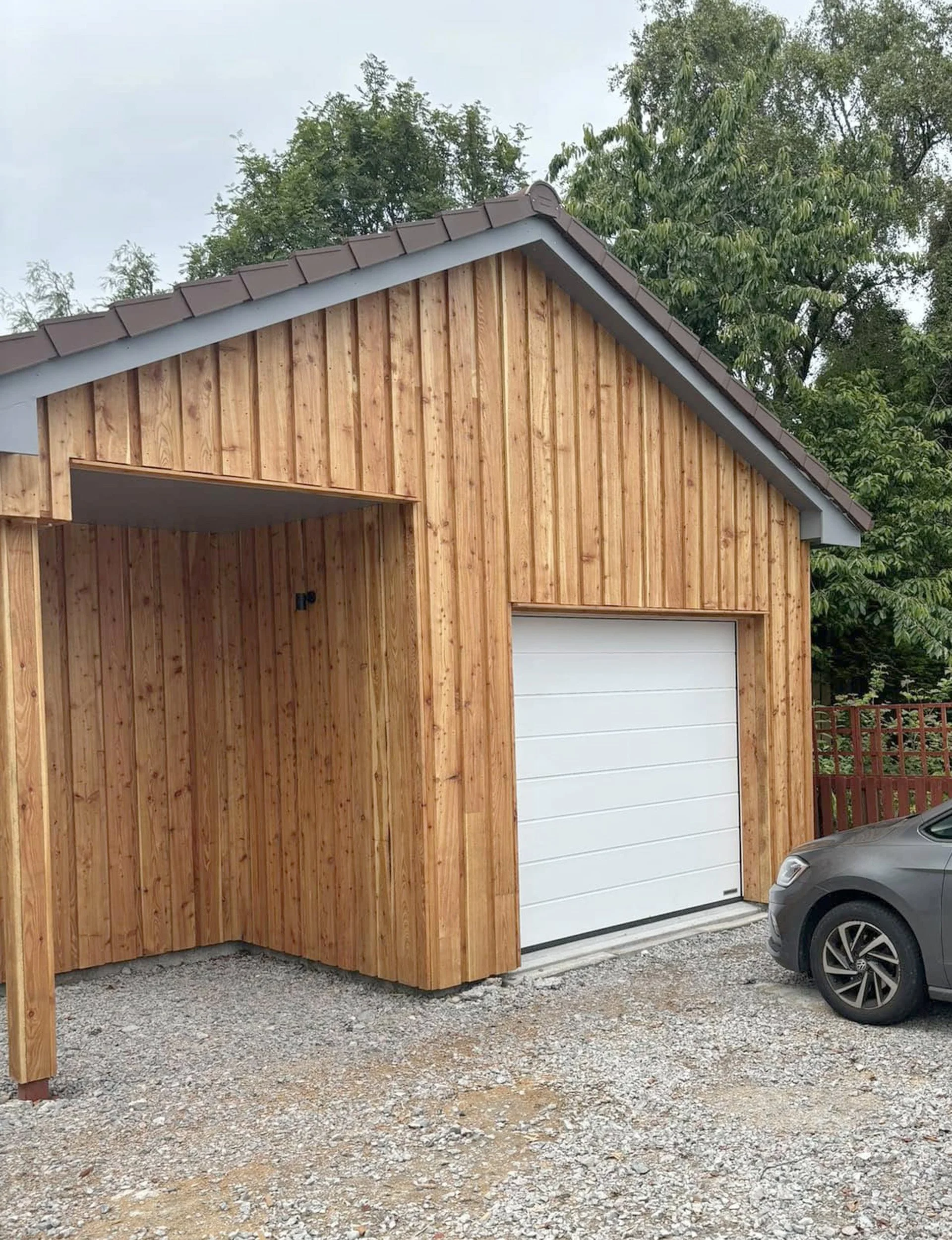 A wooden garage with a white roll-up door, adjacent to a small car, on a gravel driveway with trees in the background.