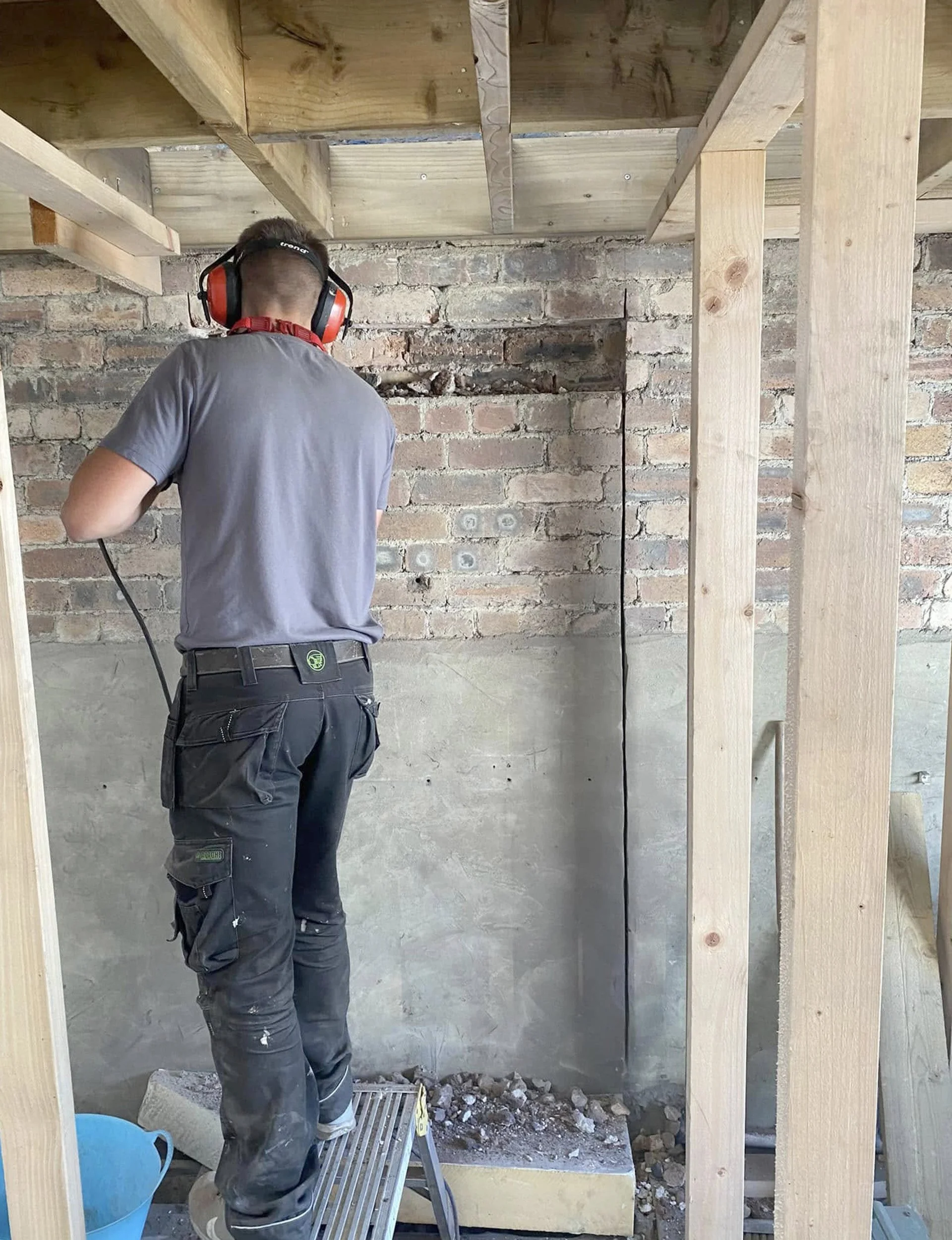 A person wearing ear protection and work clothes standing on a step ladder, working on a brick and concrete wall in a construction site.