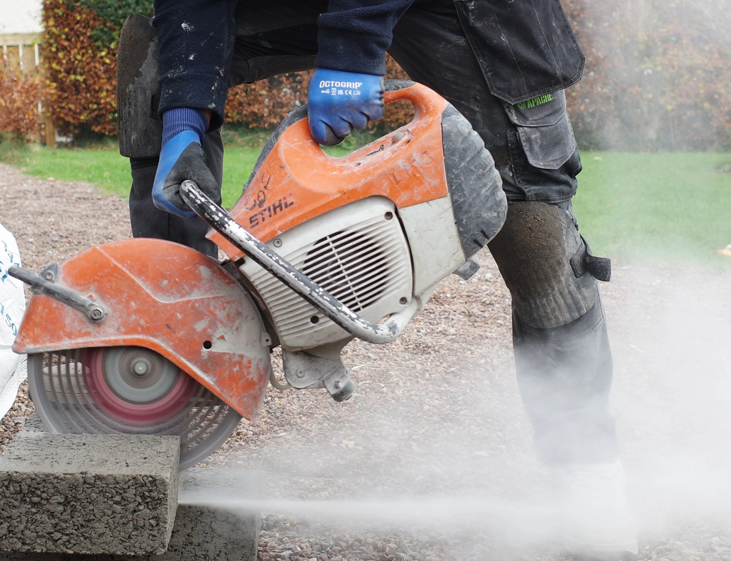 A person using a handheld concrete cutter on a stone block outdoors, wearing gloves and protective clothing.