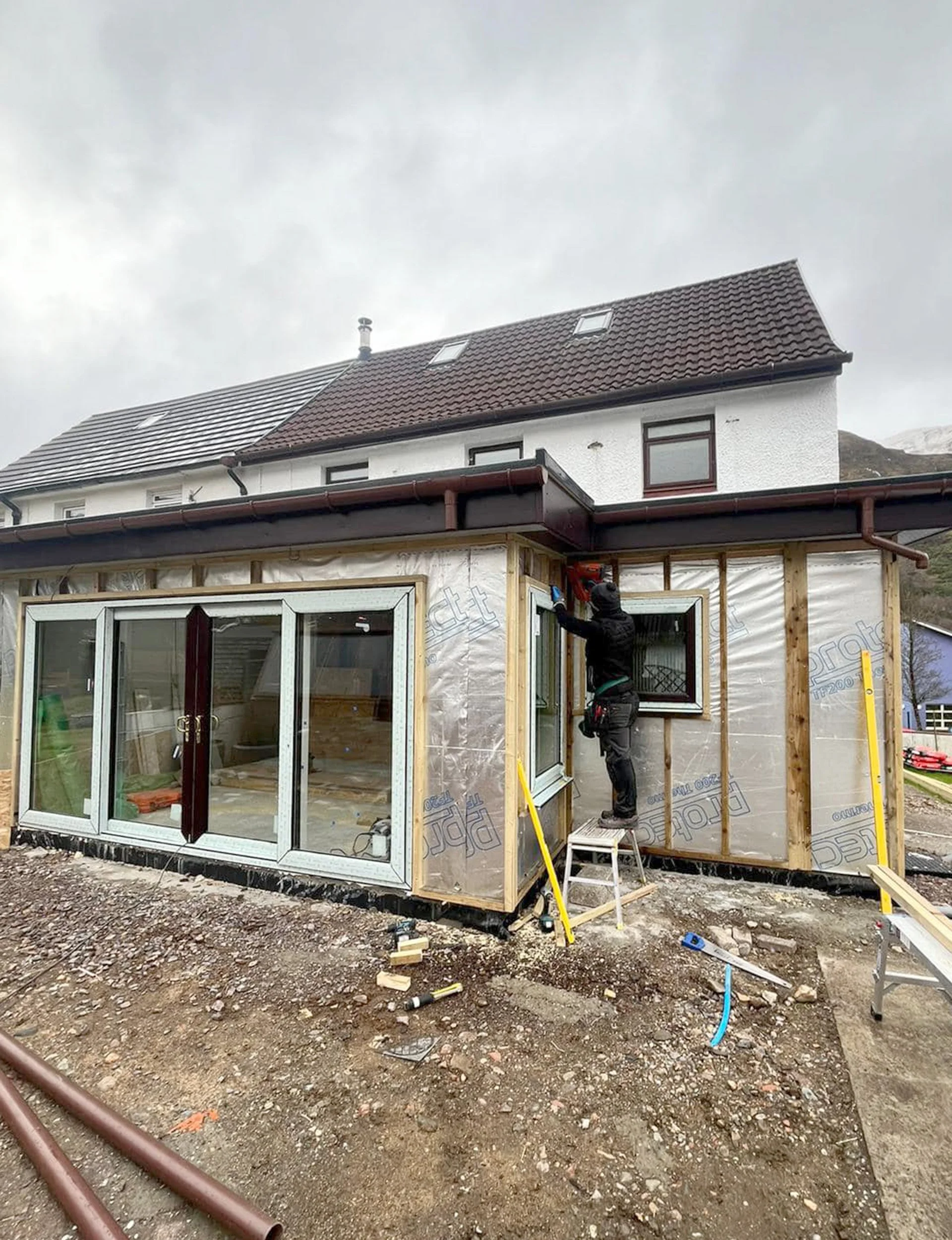 A construction worker installing a window on the exterior of a house during renovation, with building materials and tools around.