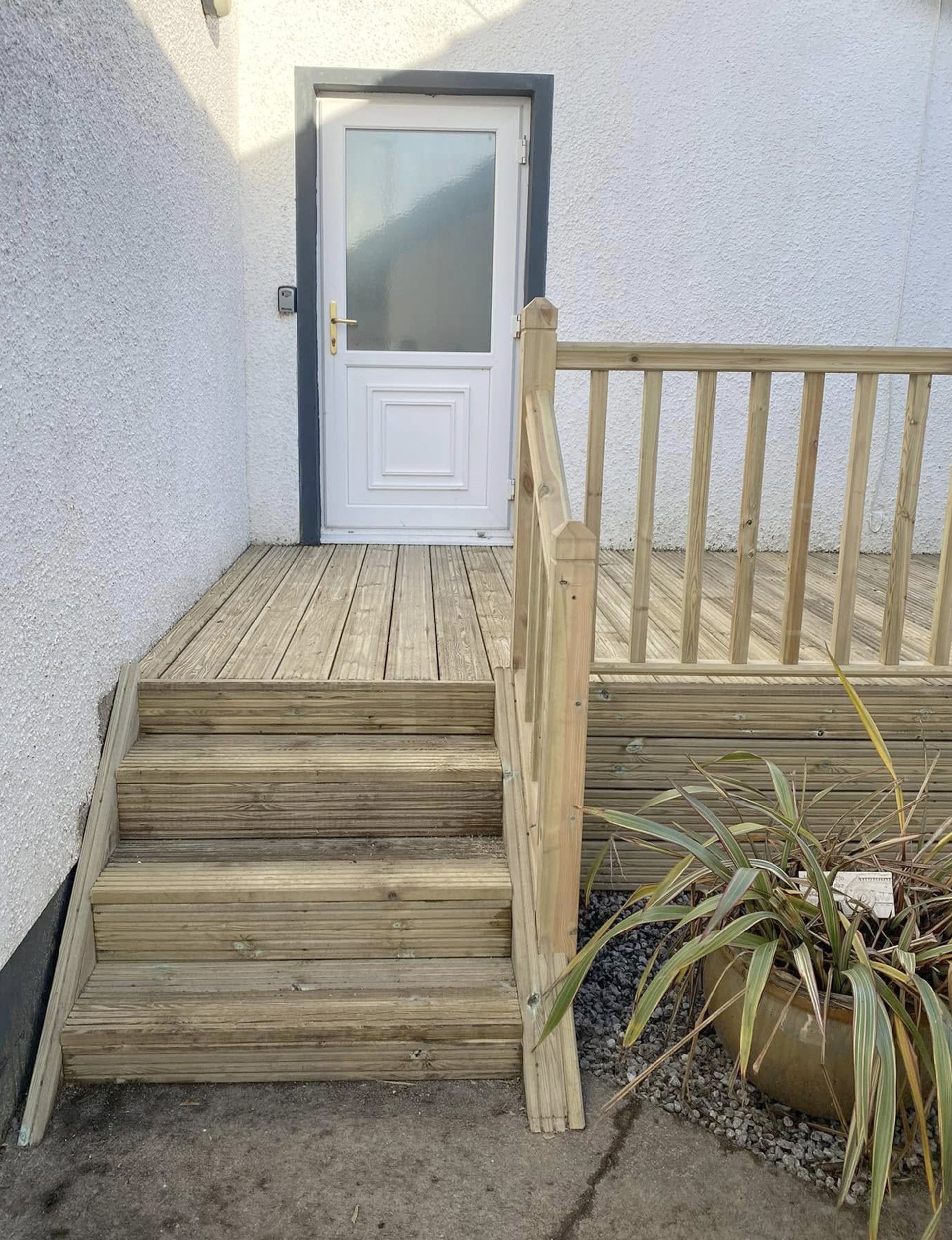 New wooden ramp and stairs leading to a white door with a frosted glass panel, attached to a white stucco building, with a potted plant nearby.