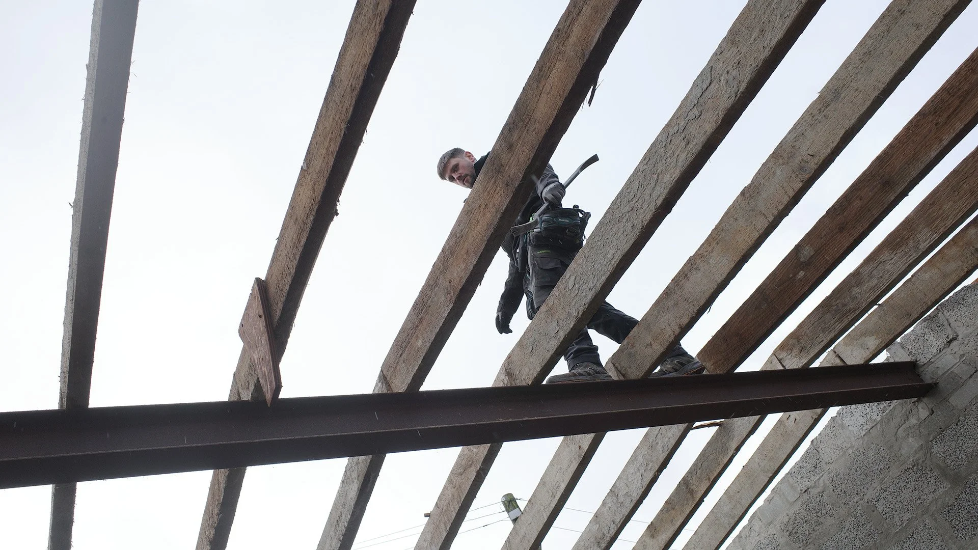 A person working on a construction site, standing on wooden beams, holding a tool, and wearing safety gear, viewed from below against a cloudy sky.