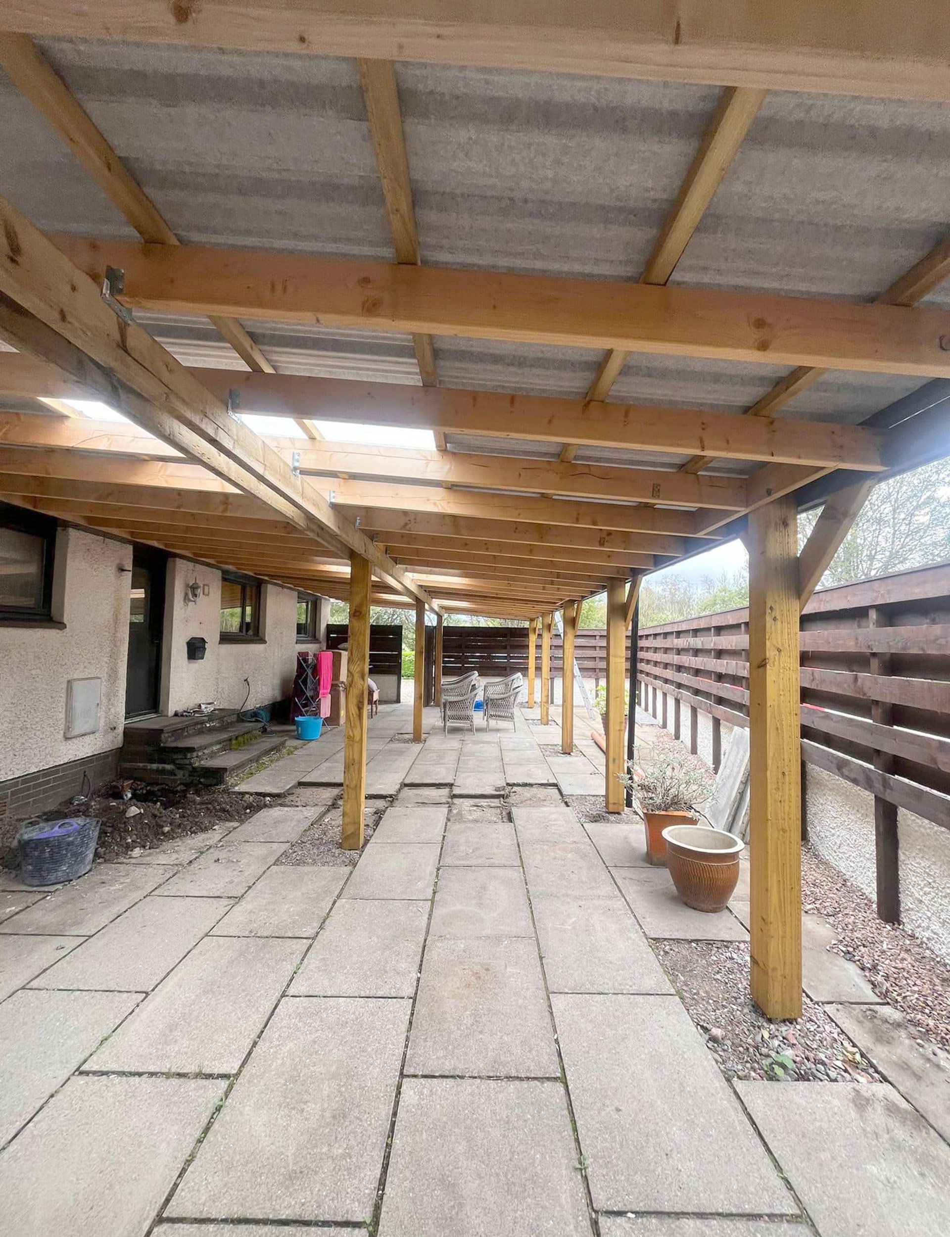 Backyard patio with a wooden porch roof supported by posts, patio furniture, potted plants, and a privacy fence.