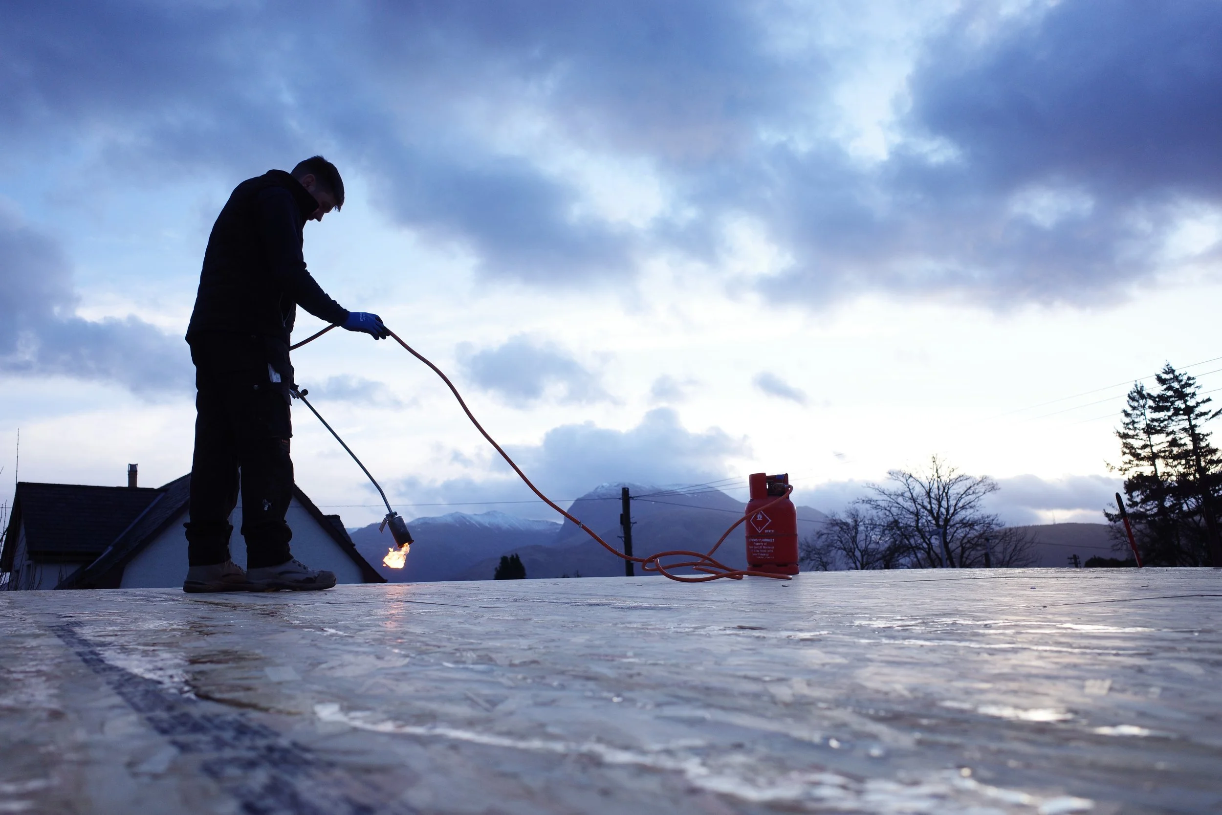 Person in silhouette working on roofing with a propane torch, with mountains and trees in the background.