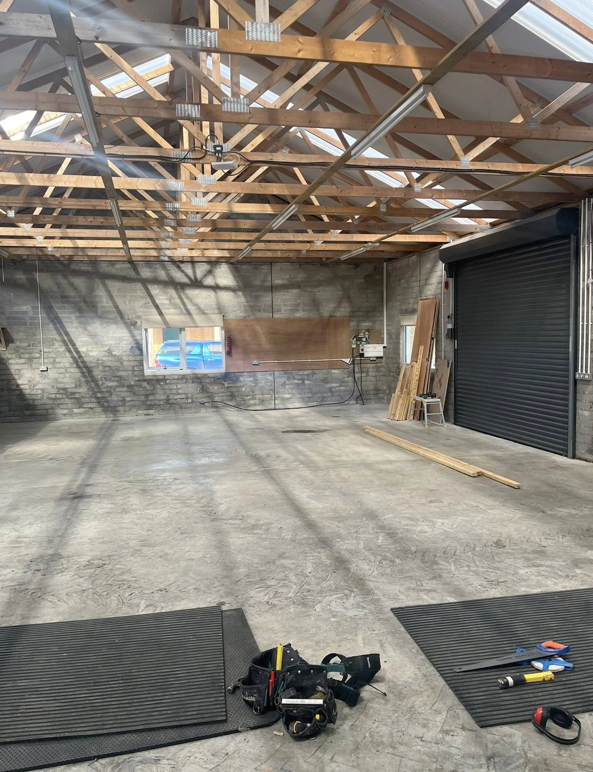 An unfinished garage or workshop space with a concrete floor, exposed wooden roof trusses, a small window, and a black roll-up door. There are tools and materials on the floor, including two black mats, a yellow utility knife, a red and black flashli