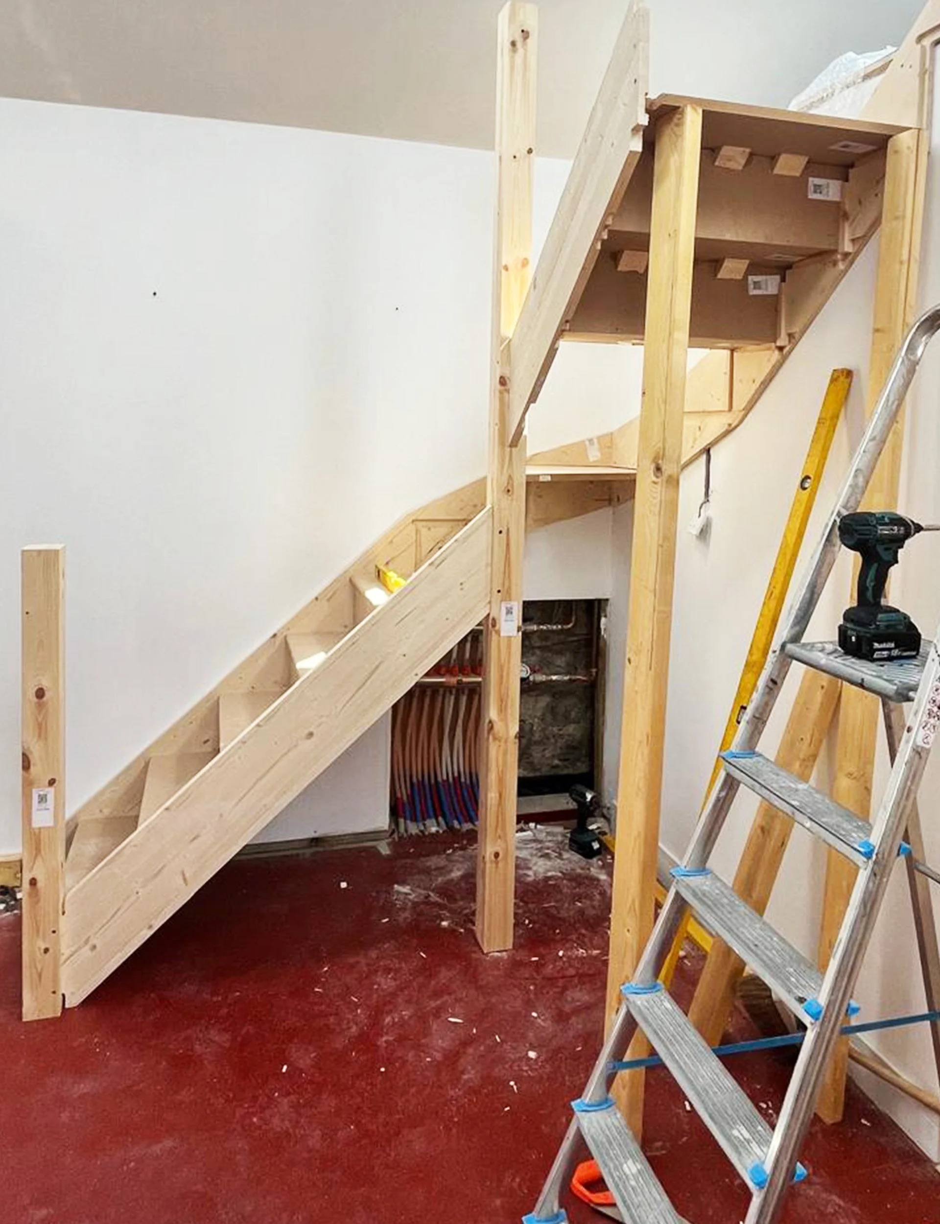 Wooden staircase under construction in a room with a red floor, with tools and a ladder nearby.