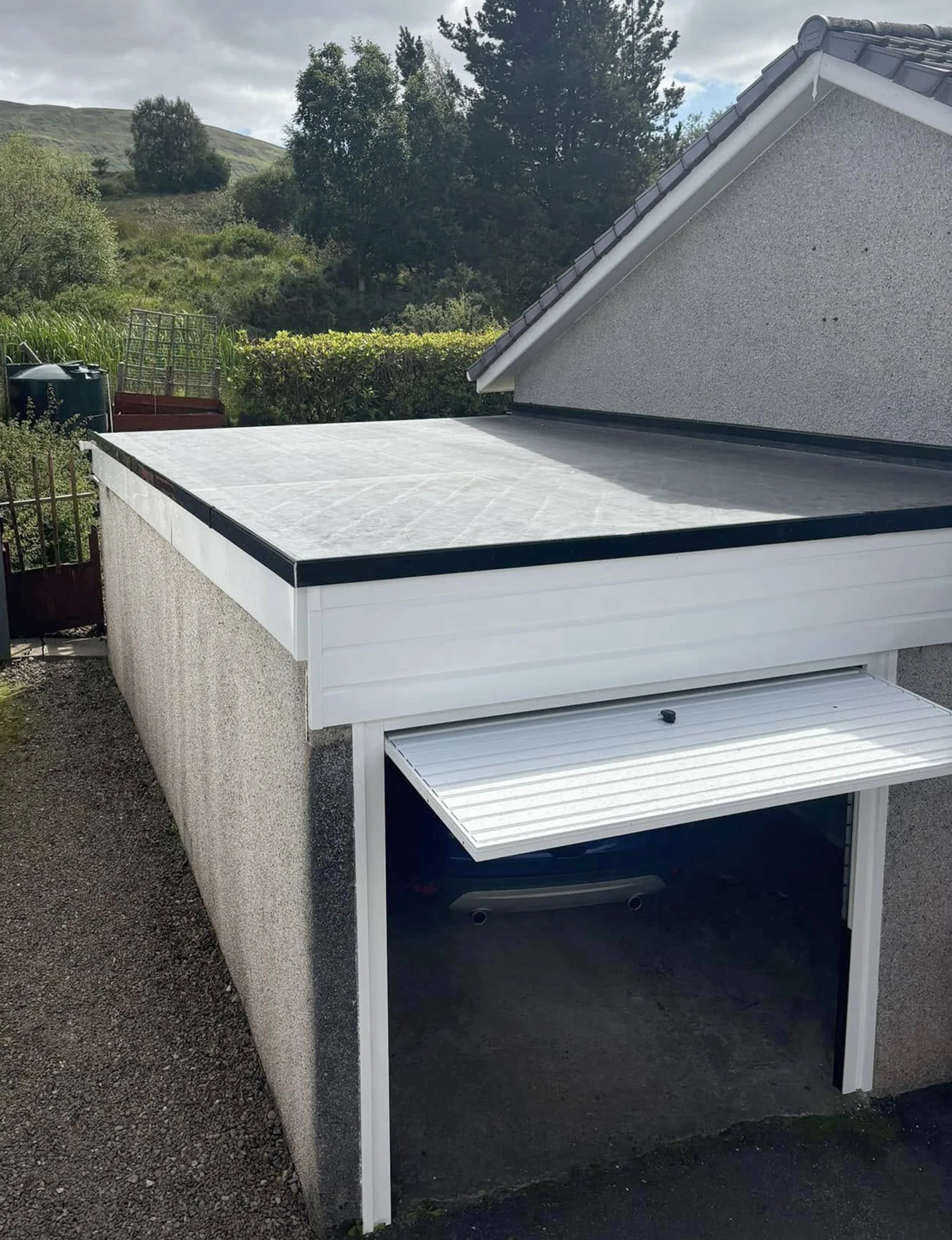 Exterior view of a residential garage with a closed white roller door, adjacent to a grey house wall, with a concrete driveway and a black trim roof, surrounded by greenery and hills in the background.