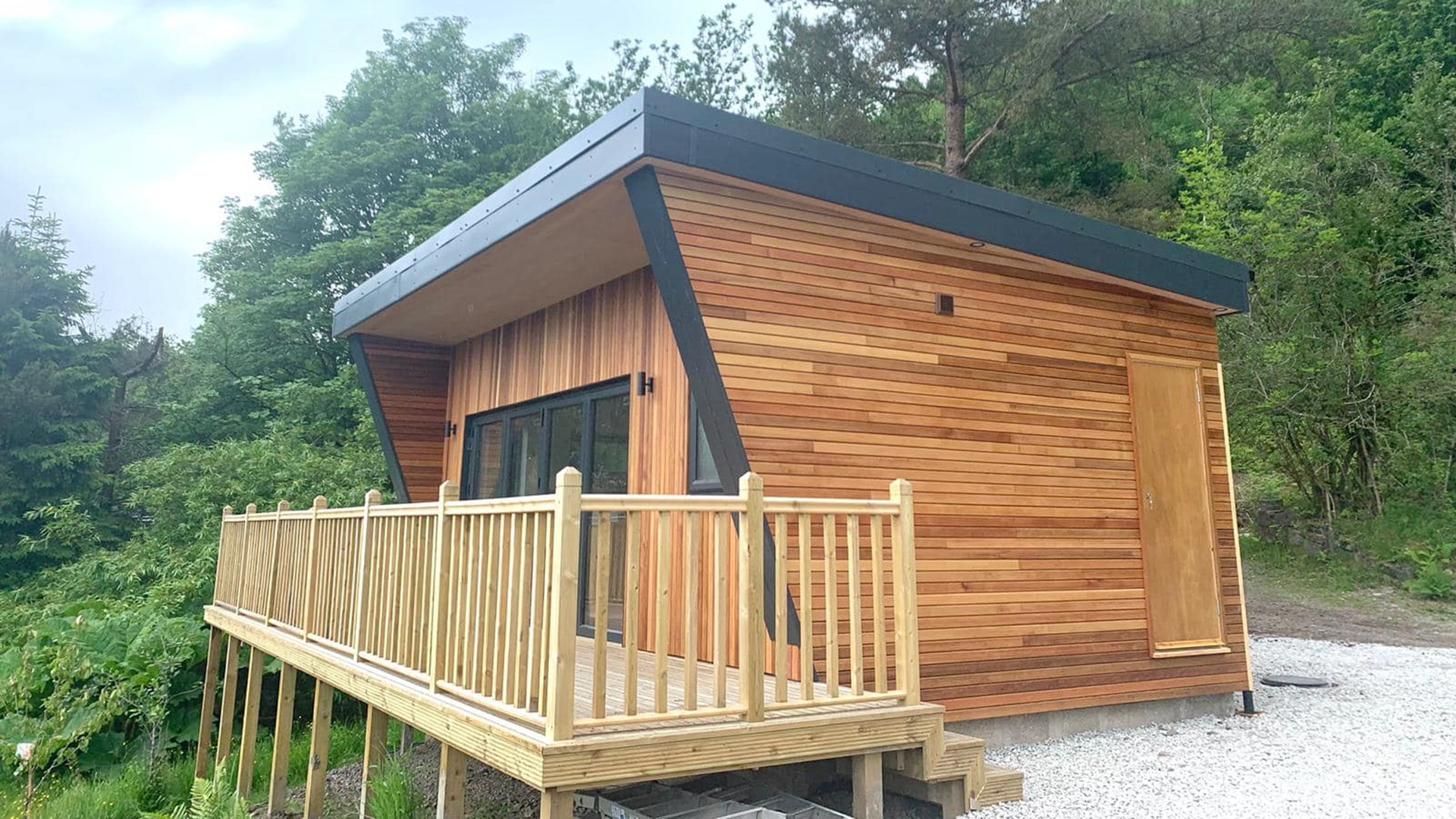 A modern wooden house with a slanted roof, located in a green, wooded area, with a small raised porch and gravel ground.