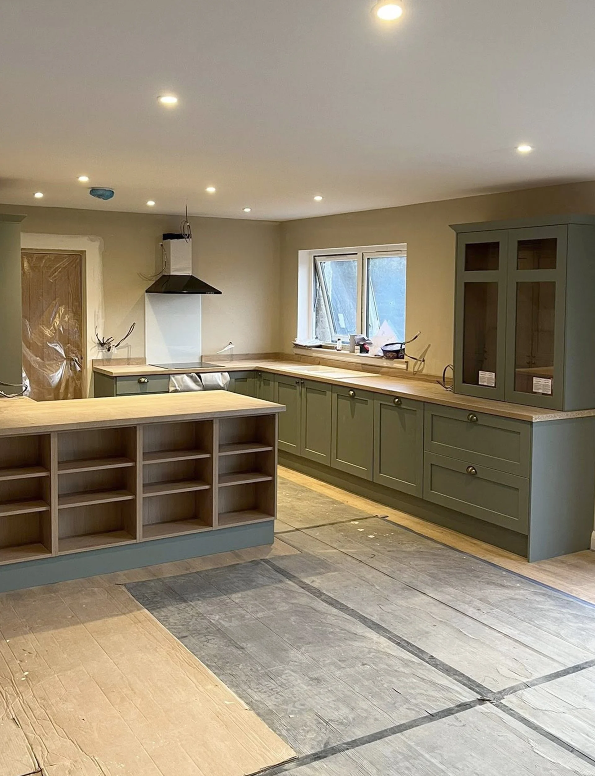 Kitchen under construction with green cabinetry, wooden countertops, an island with open shelves, and partially finished flooring.