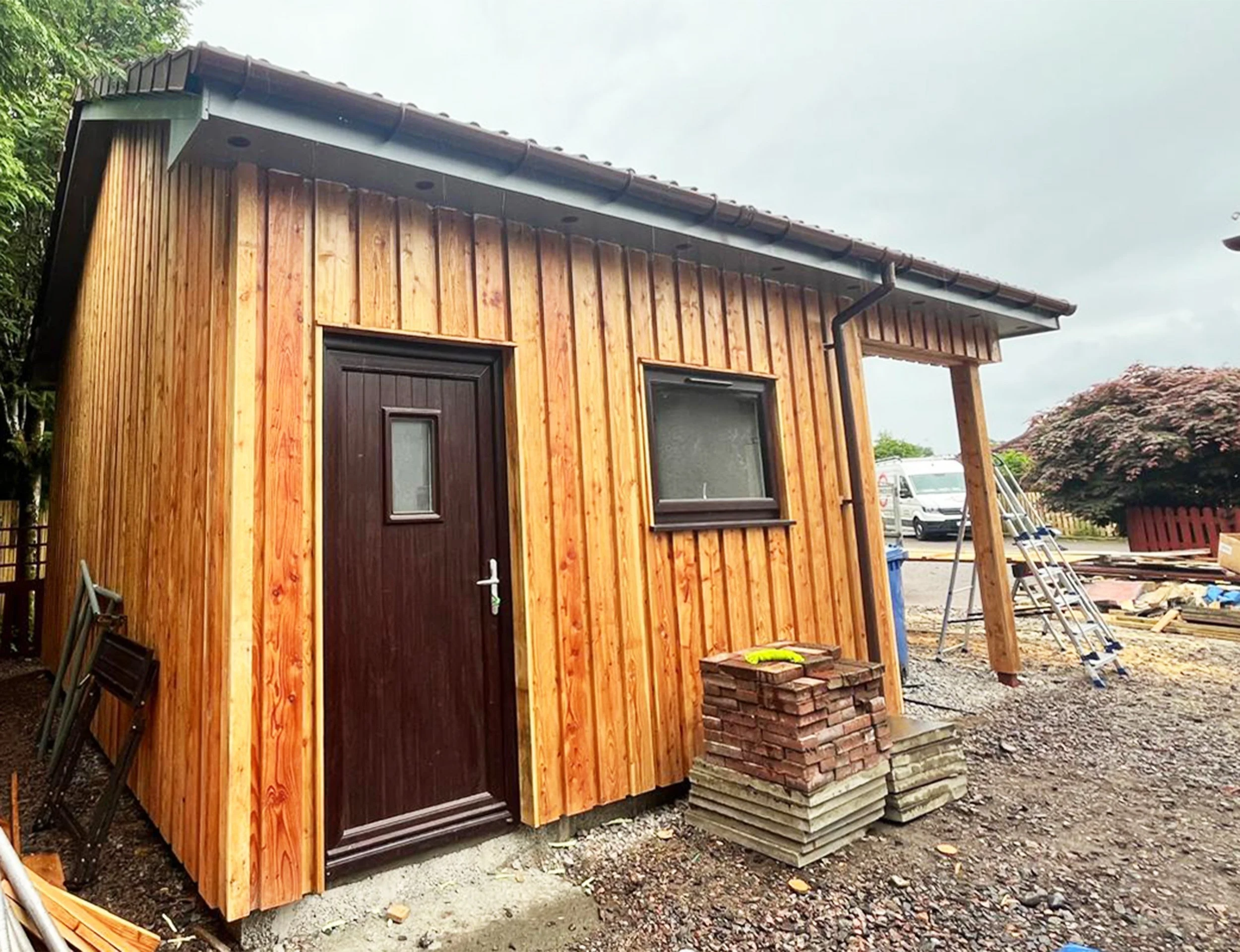 Small wooden building under construction with a dark brown door, a window, and construction materials outside. Adjacent to the building are ladders and a pile of bricks. The scene appears to be a work site with vehicles and a tree in the background.