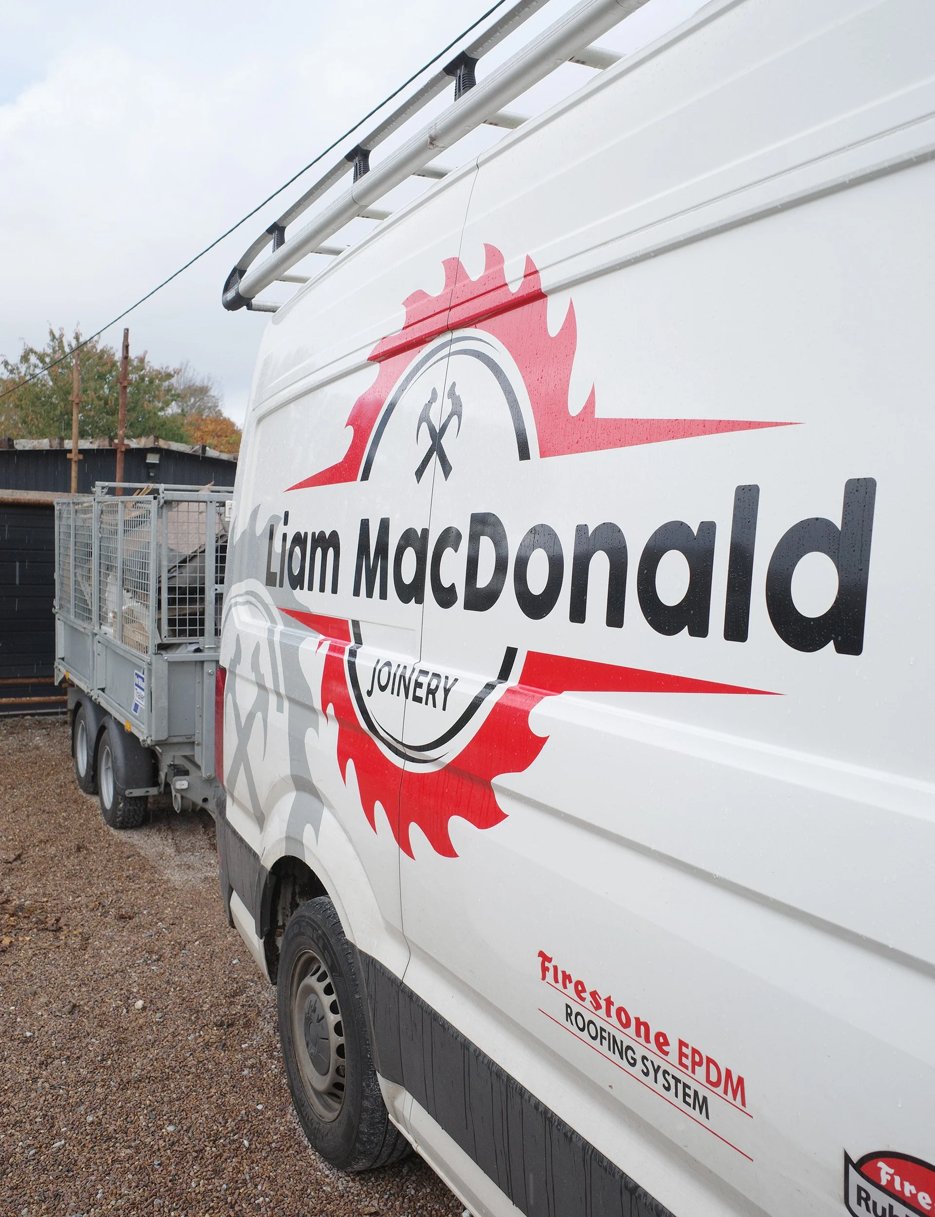 A white commercial van with the logo of Liam MacDonald Joinery, featuring a red and black saw blade design and the words Firestone EPDM Roofing System written on the side. The van is parked outdoors on gravel with a black shed or building and trees in the background.