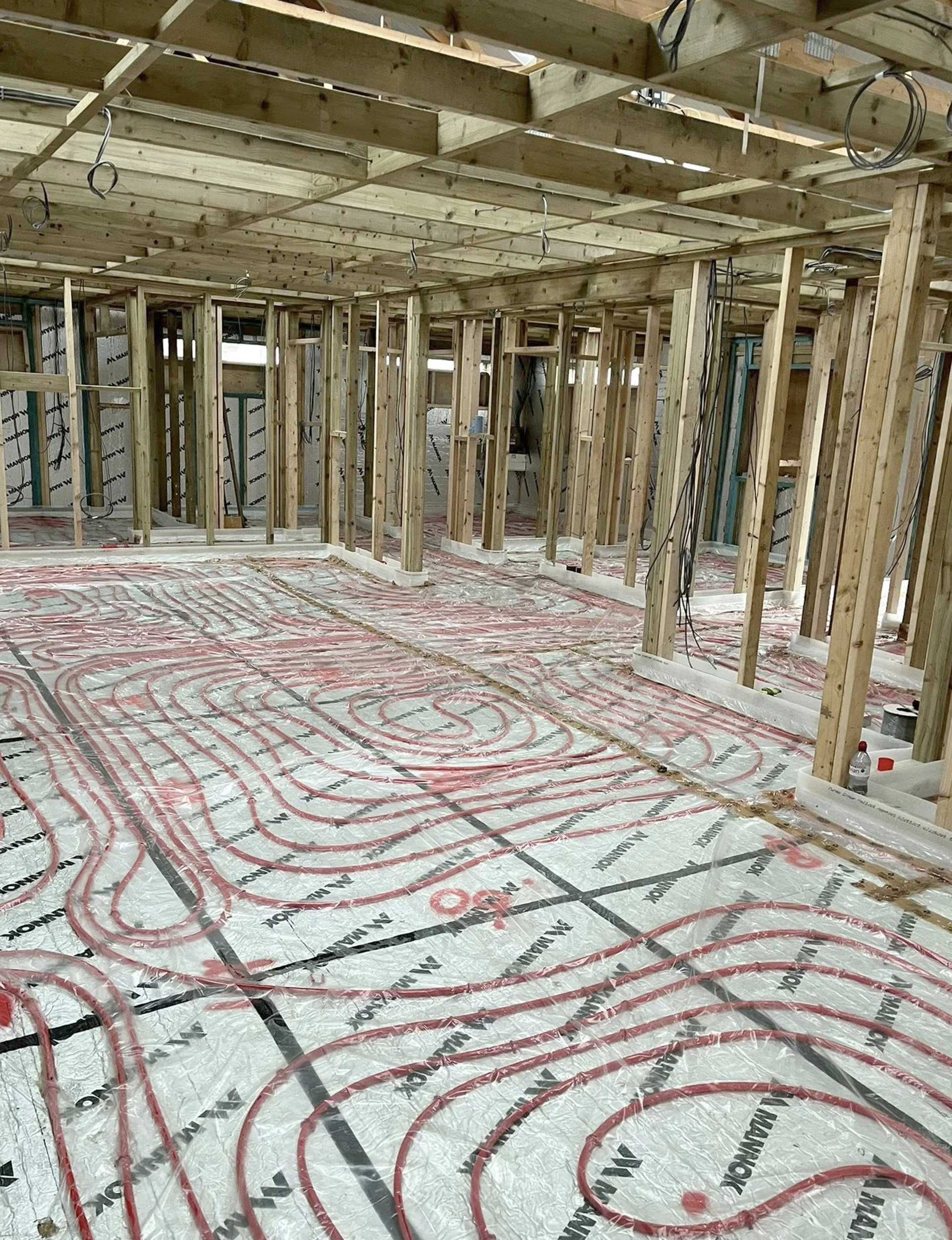 Interior view of a building under construction with wooden framing and a radiant floor heating system installed, showing red tubing on the floor.