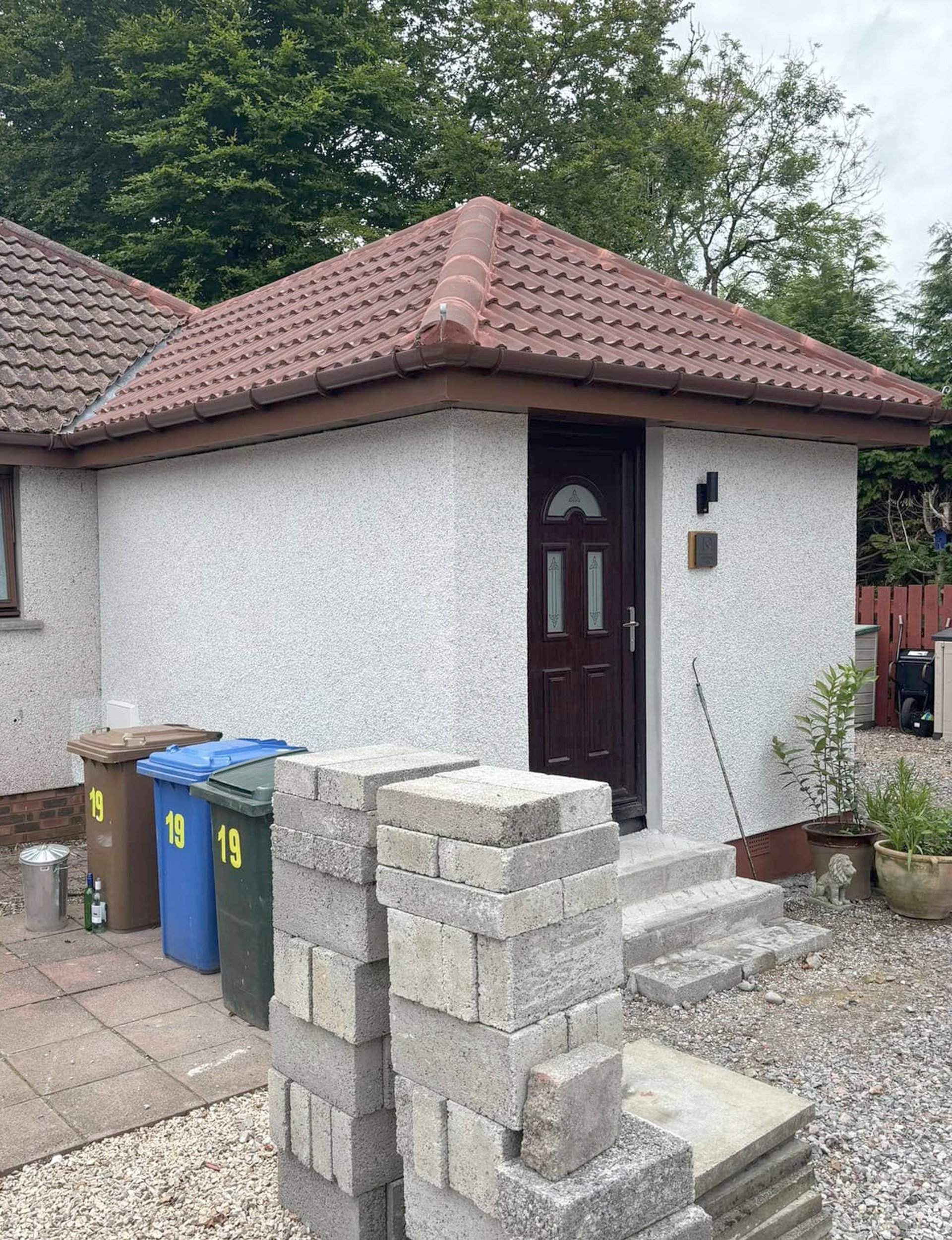 A small house with a red tiled roof, a dark wooden front door, and white exterior walls. There are three trash bins at the side of the house, with yellow number 19 on them. In front of the house, there are two small steps leading to the door, with so