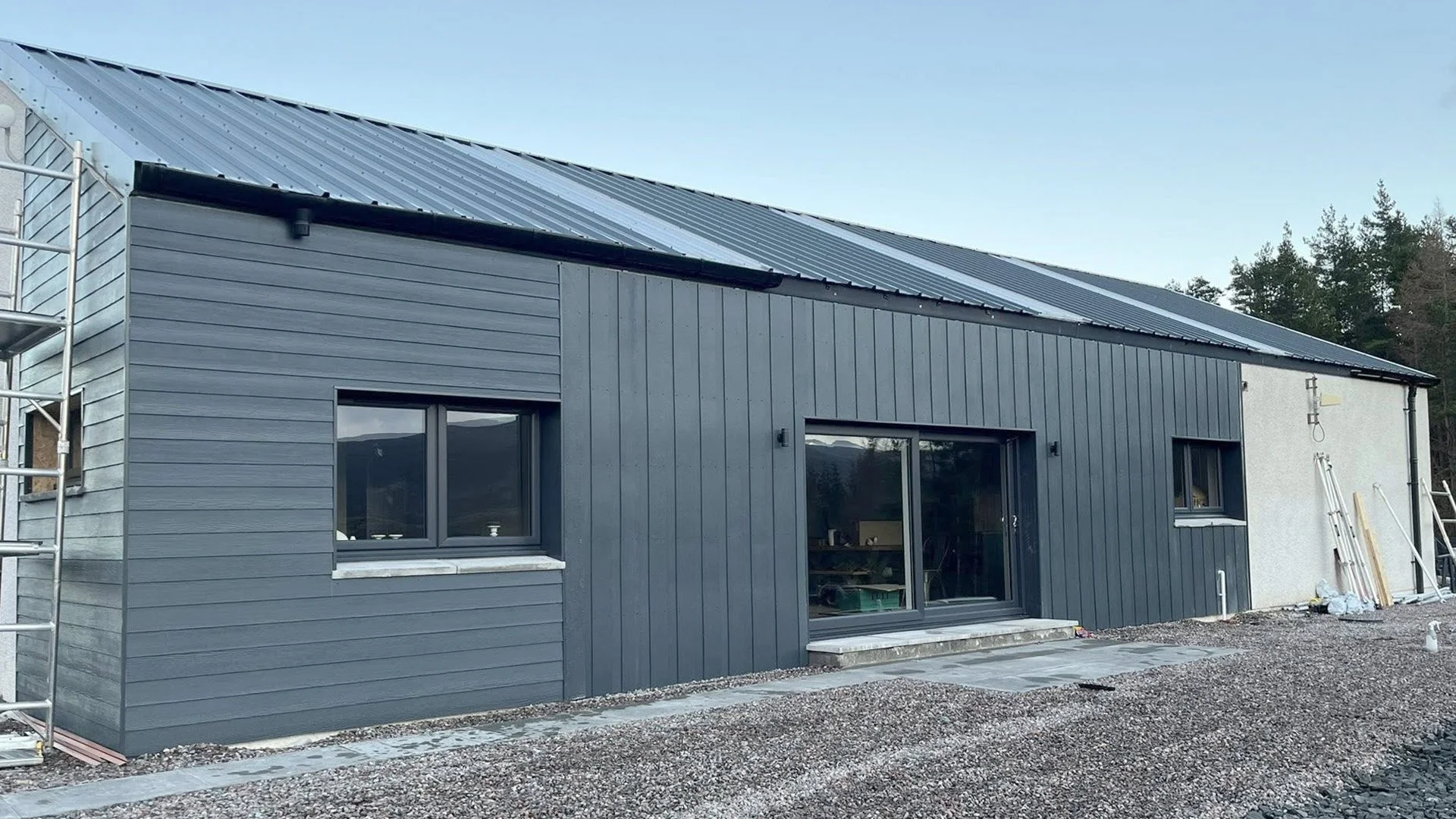 Modern house under construction with black vertical siding, a sliding glass door, and a small window, surrounded by construction materials and scaffolding, with trees in the background.