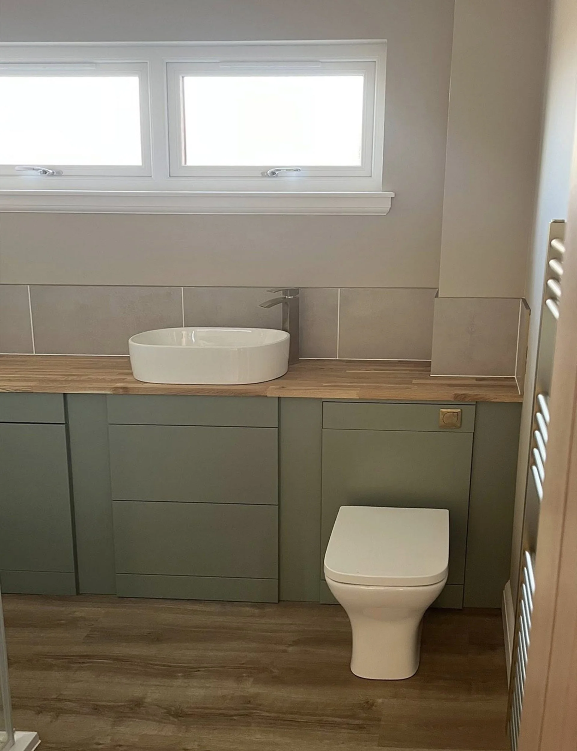 A small bathroom with a green vanity, wooden countertop, vessel sink, silver faucet, white toilet, two rectangular window panes, beige tiled backsplash, and wooden floor.