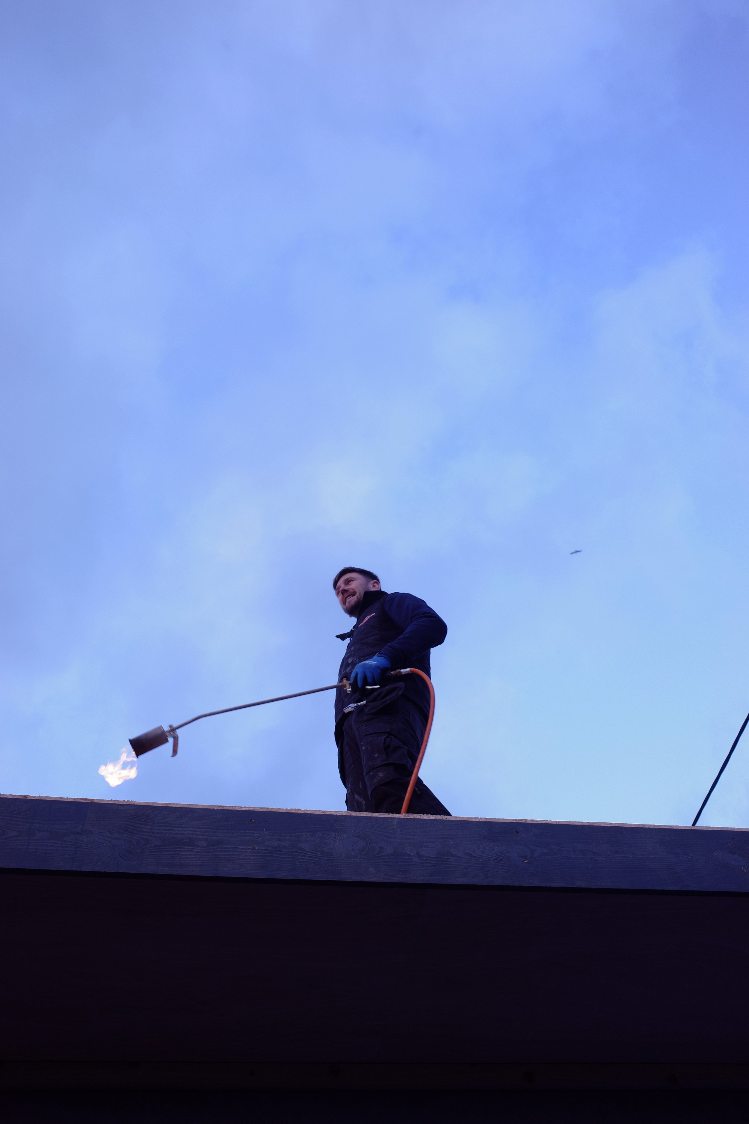 A man standing on a rooftop holding a blowtorch with a flame, using safety gloves, with a clear blue sky in the background.