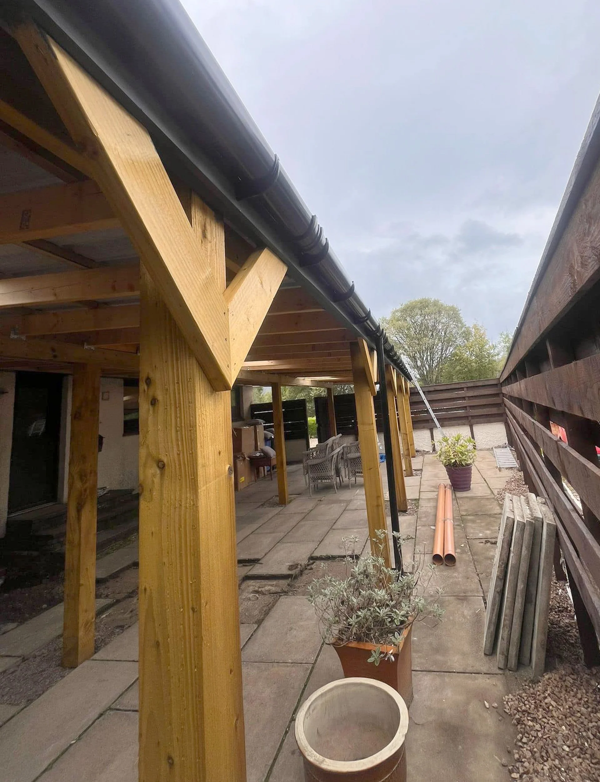 Back patio area with wooden pergola under construction, black rain gutter, potted plants, outdoor furniture, and construction materials like pipes and wood planks.