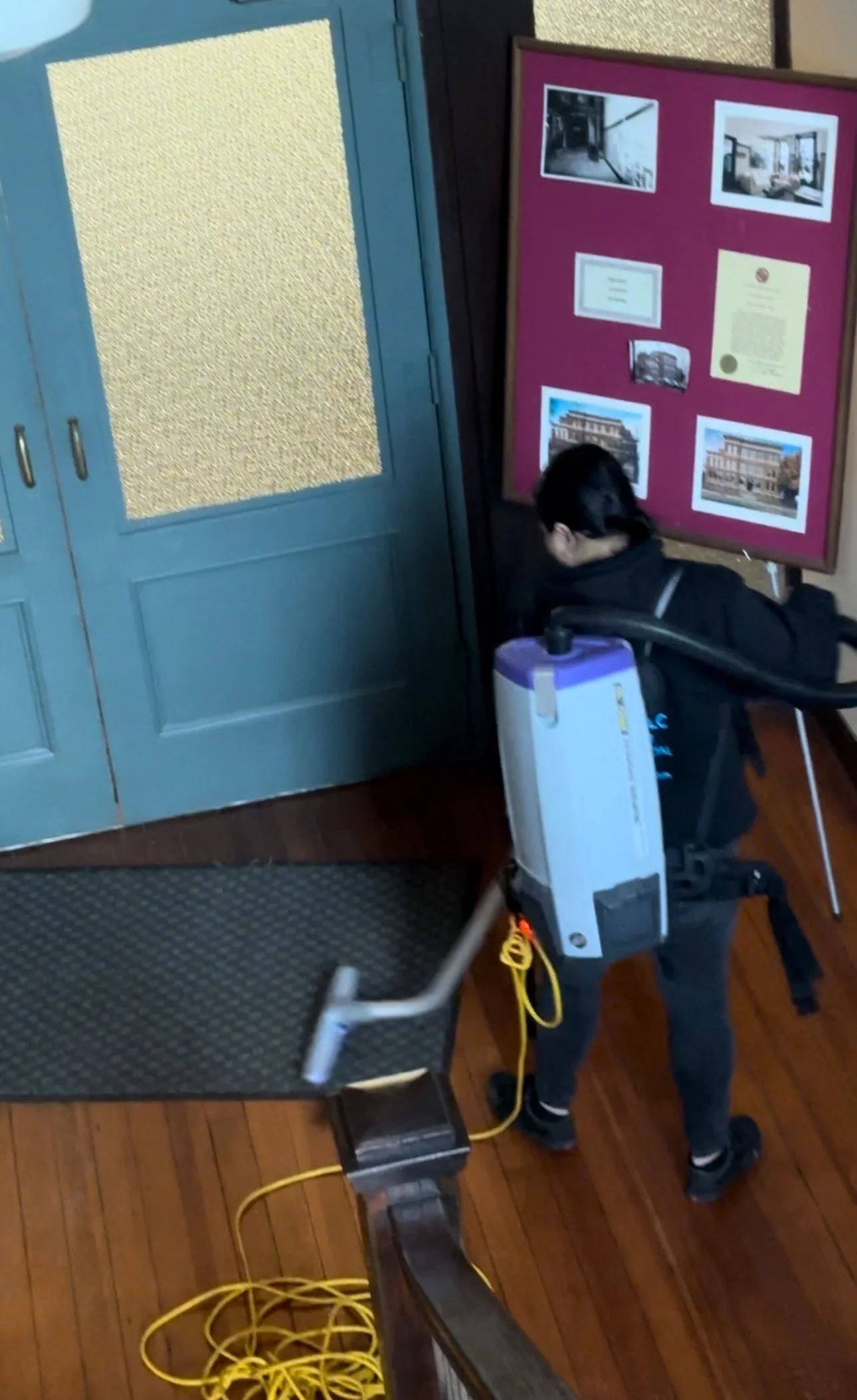 Person in a wheelchair beside a blue door and framed historical display, with a portable oxygen concentrator and yellow cable.