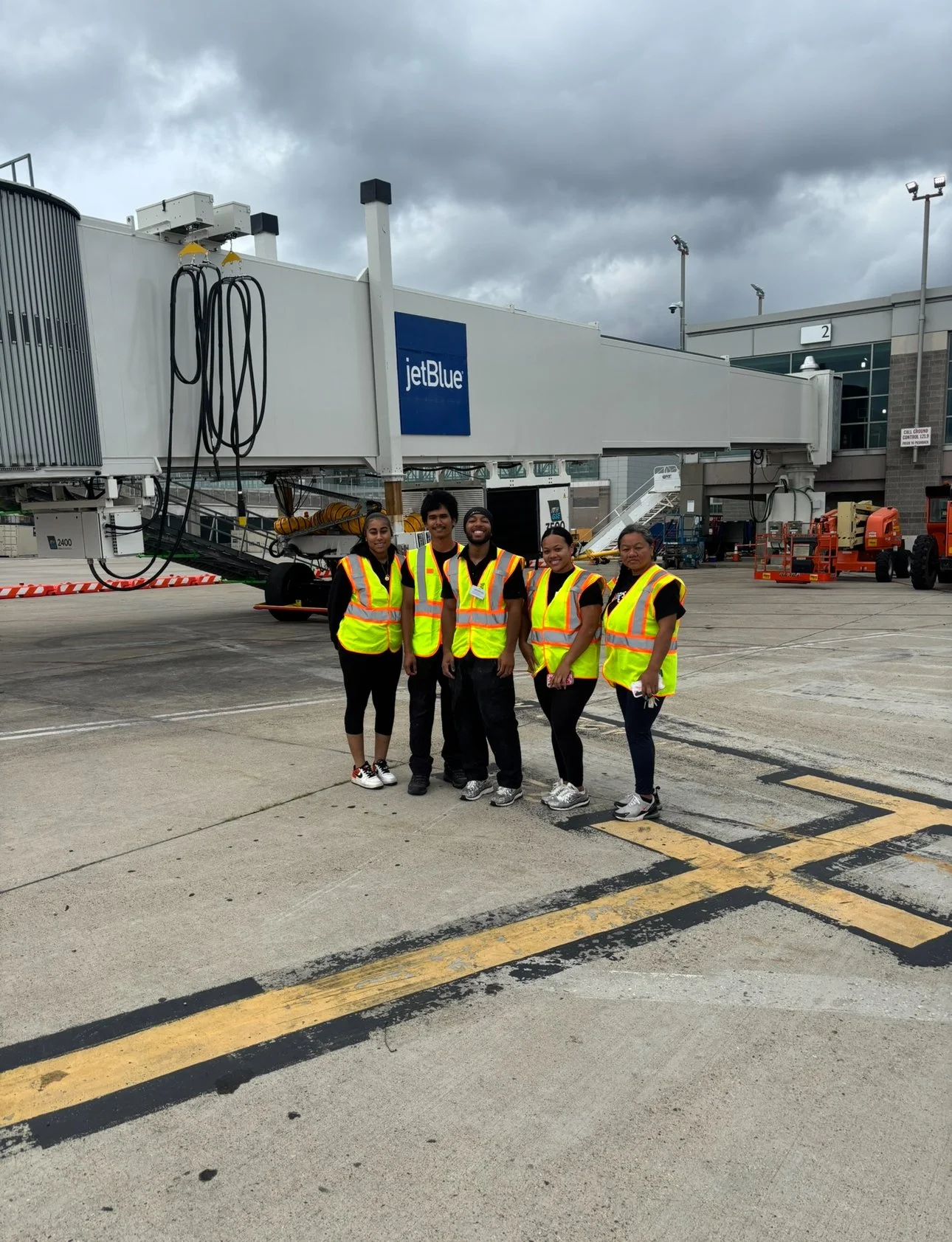 Five airport workers in high-visibility vests pose outside a JetBlue gate beneath a cloudy sky.