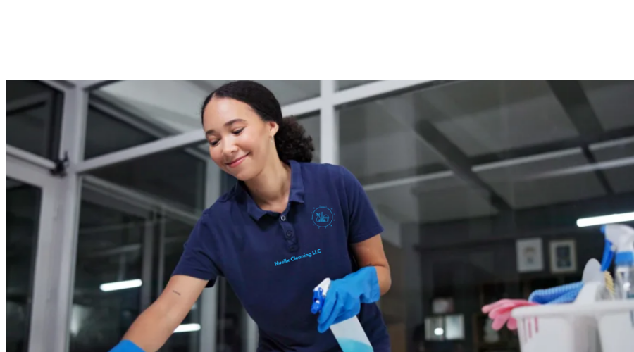 A woman wearing a navy blue polo shirt and blue gloves cleaning a surface with a spray bottle in an office or commercial space.