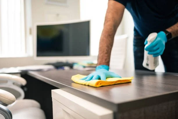 Person wearing blue gloves cleaning a dark brown desk with a yellow cloth and spray bottle, with a computer monitor and office chairs in the background.