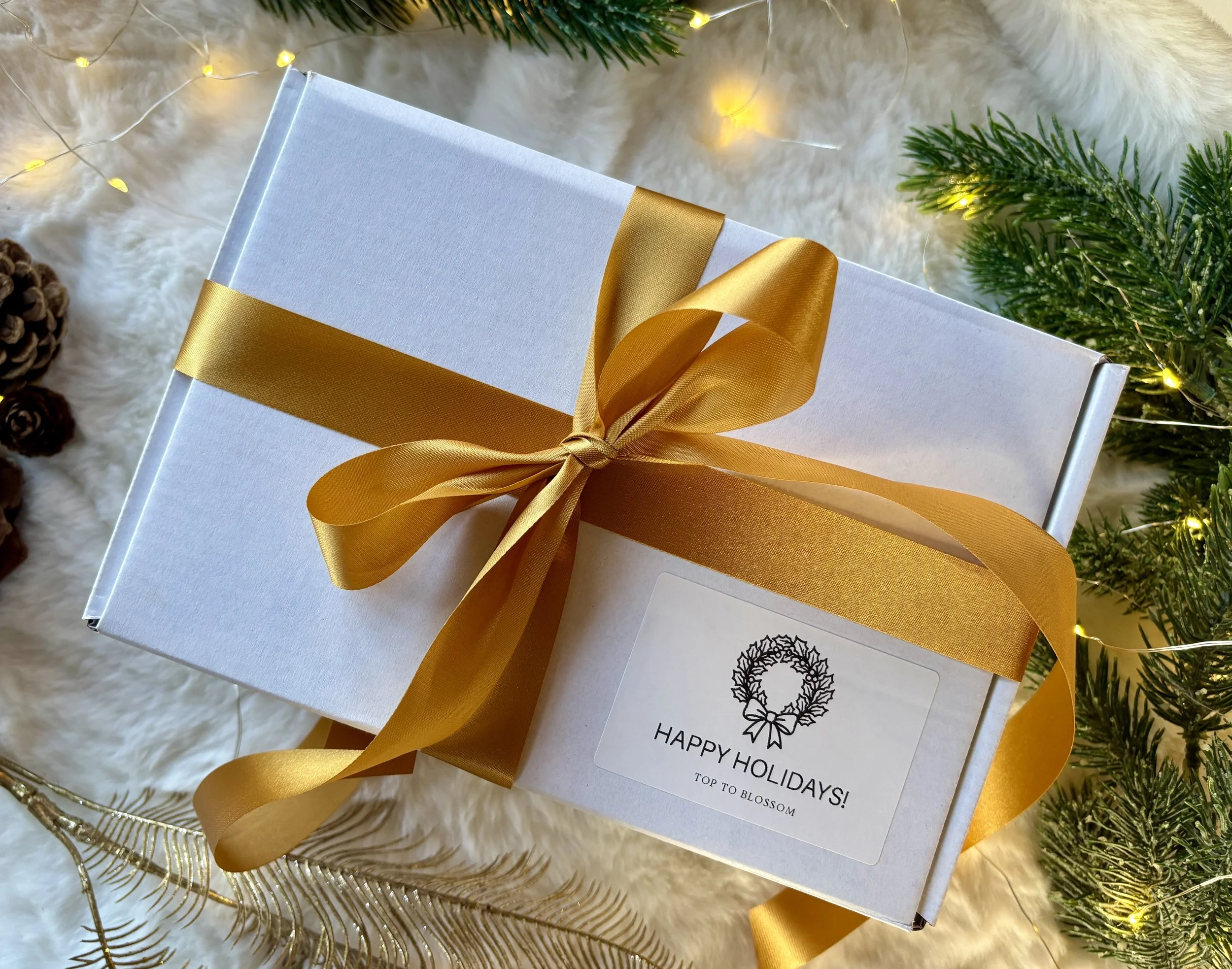 Wit cadeau met een gouden strik en een label met de tekst 'Happy Holidays! Top to Blossom', gelegen tussen kerstdecoraties zoals dennenboompjes, gouden takken en lichtjes. Gevuld met natuurlijke en handgemaakte zelfverzorgingsproducten.