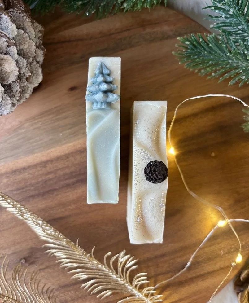 Twee botanische zeep bars met kerstthema, een met een blauw kerstboompje en de andere met zilveren ecologische glitter, omgeven door kerstdecoraties en lichtjes.