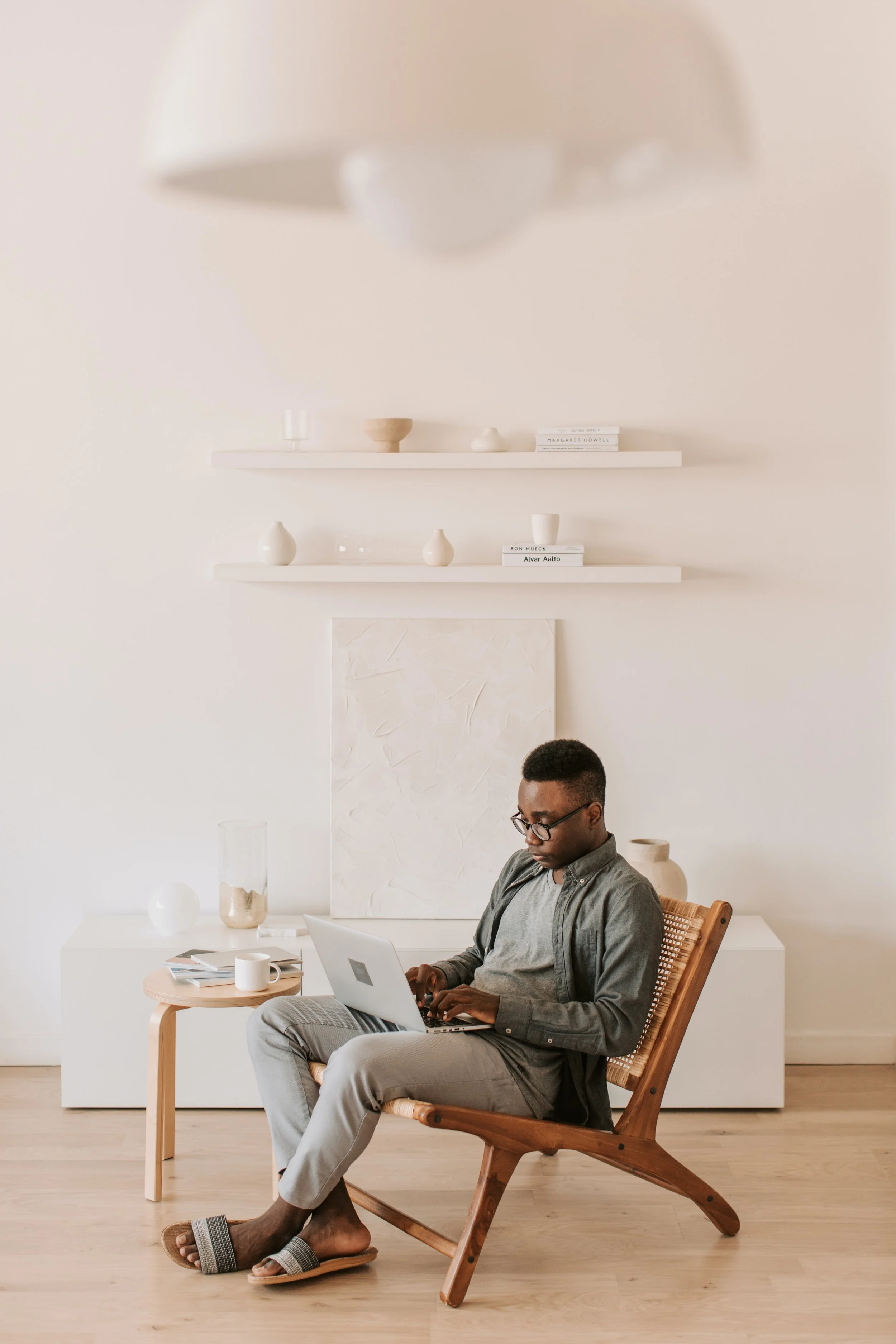A man sitting in a wooden chair working on a laptop in a minimalist room with neutral-colored decor, shelves with vases, books, and art.