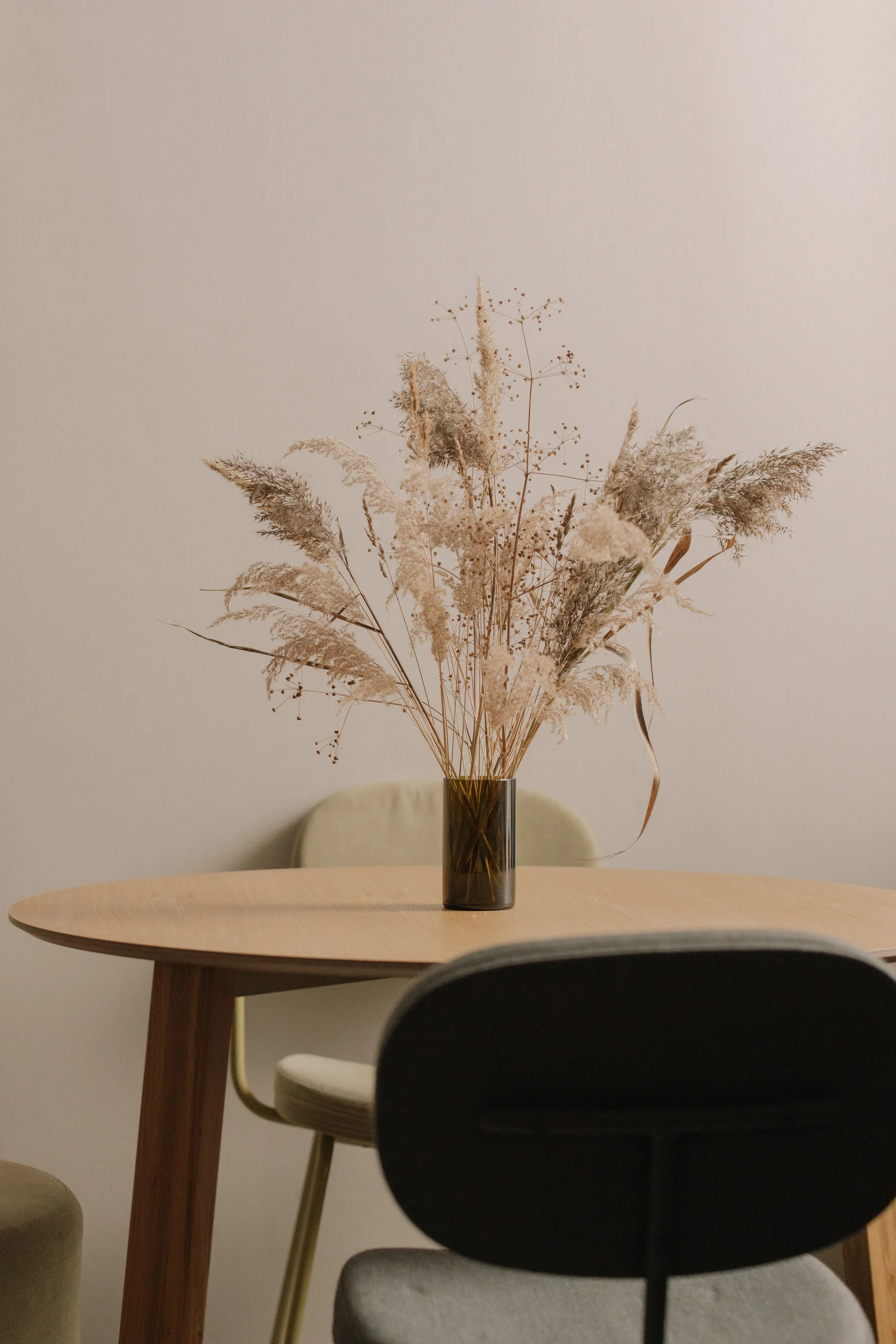 A vase with dried pampas grass and other dried flowers on a wooden table, with a chair in the foreground and a plain wall in the background.