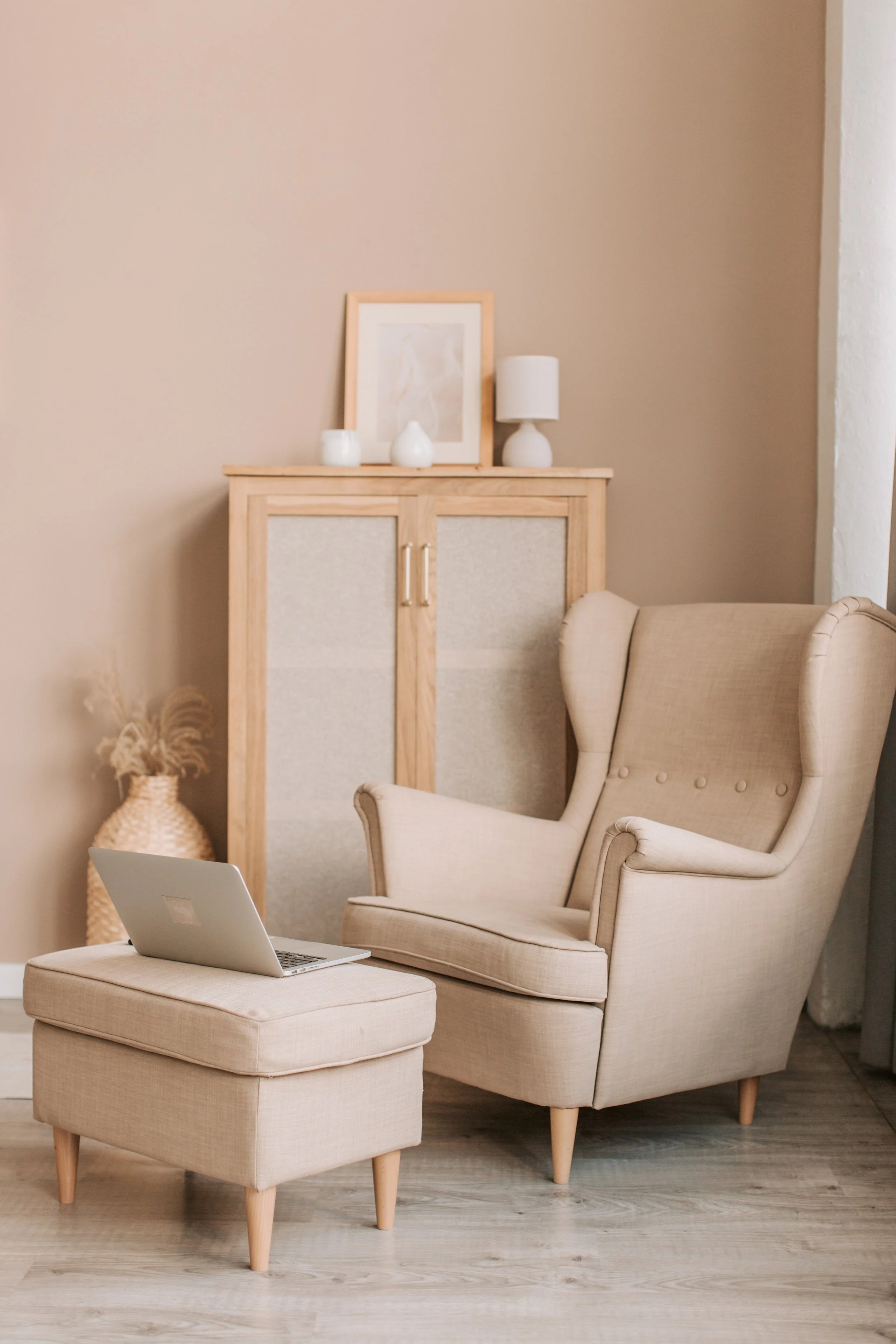A cozy reading nook with a beige high-back armchair, a matching ottoman with a laptop on top, a wooden cabinet with a framed photo, a white table lamp, vases, and a woven basket with dried flowers, all in neutral tones.