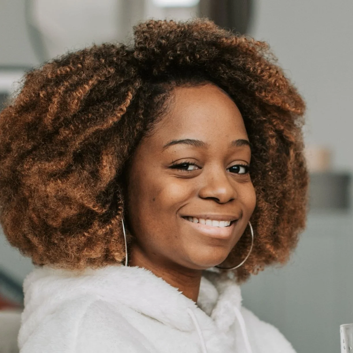 A young woman with curly, reddish-brown hair smiling and wearing hoop earrings and a white hoodie.
