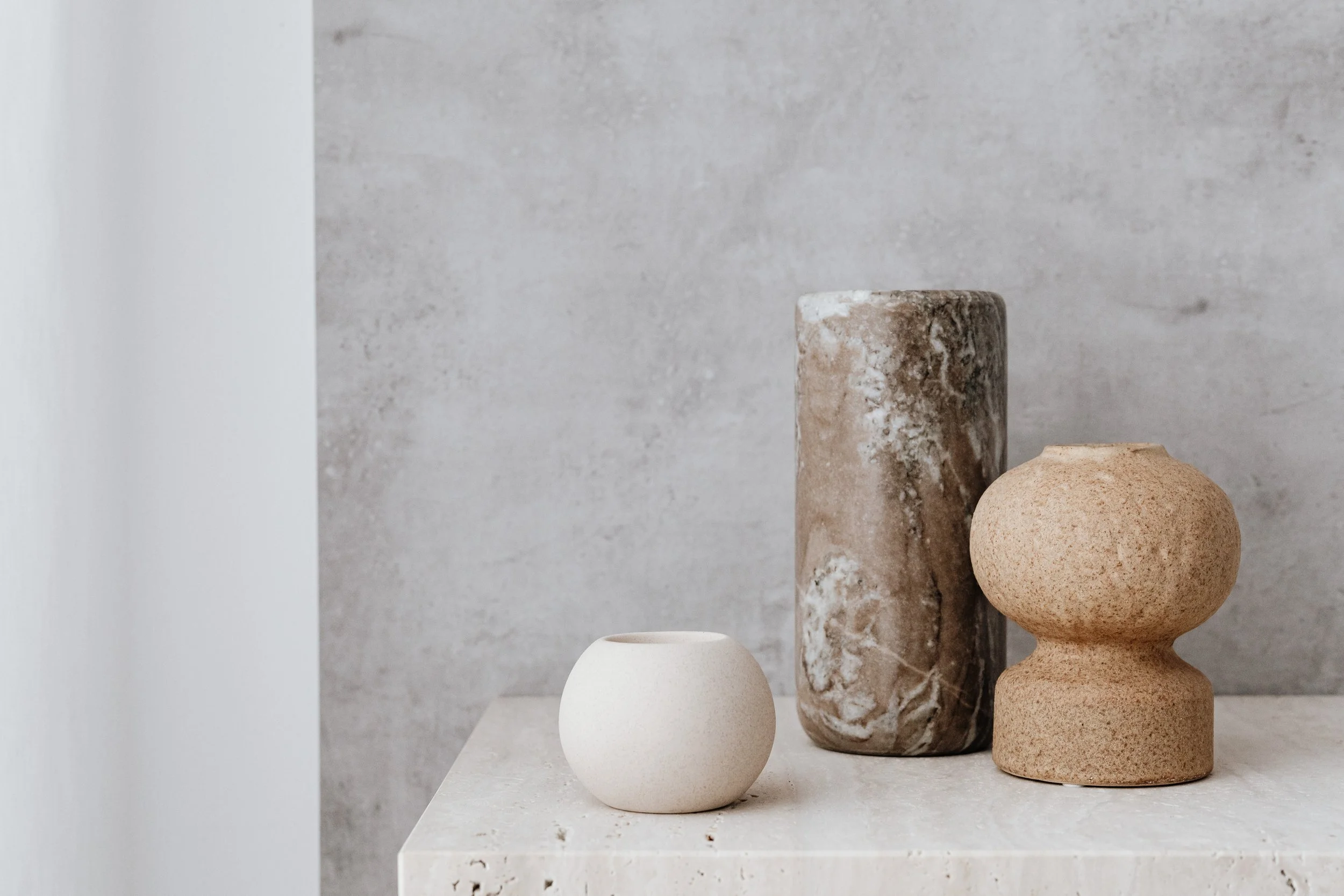 Three decorative vases of different sizes and textures on a white surface against a gray wall. The smallest is a smooth, round white vase; the middle is a tall, cylindrical brown marble vase; the largest is a textured, beige double-bulb shaped vase.