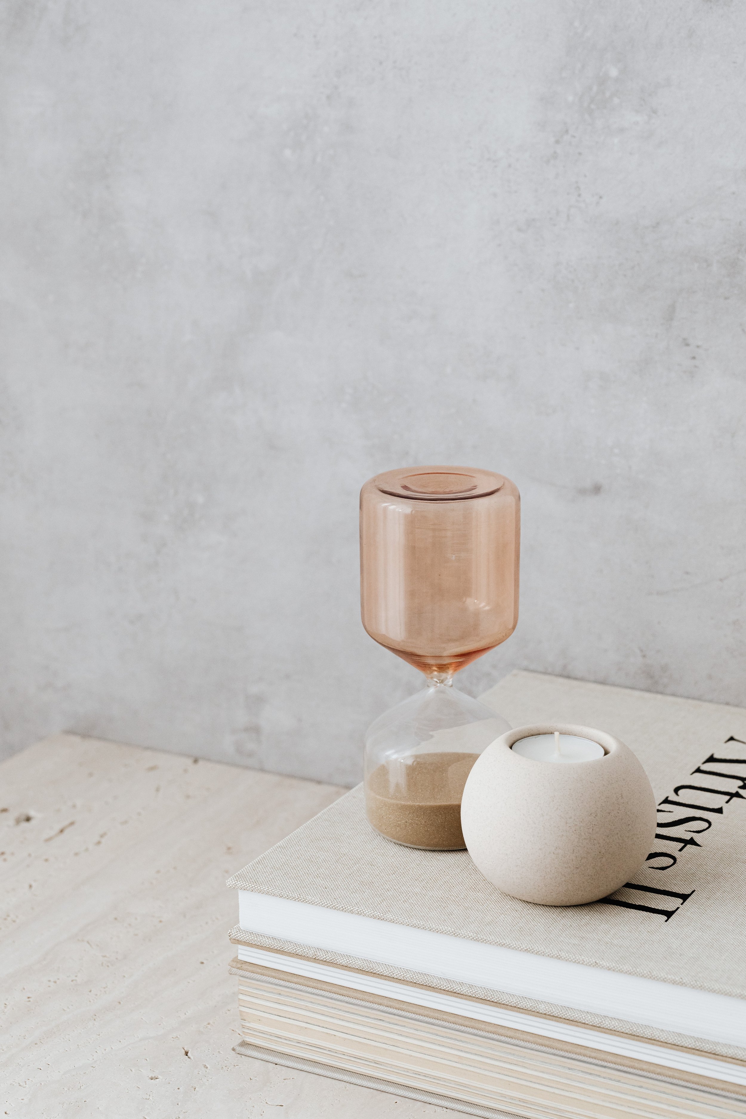 Decorative items including an hourglass and a candle holder placed on a closed book on a light-colored table against a textured gray wall.
