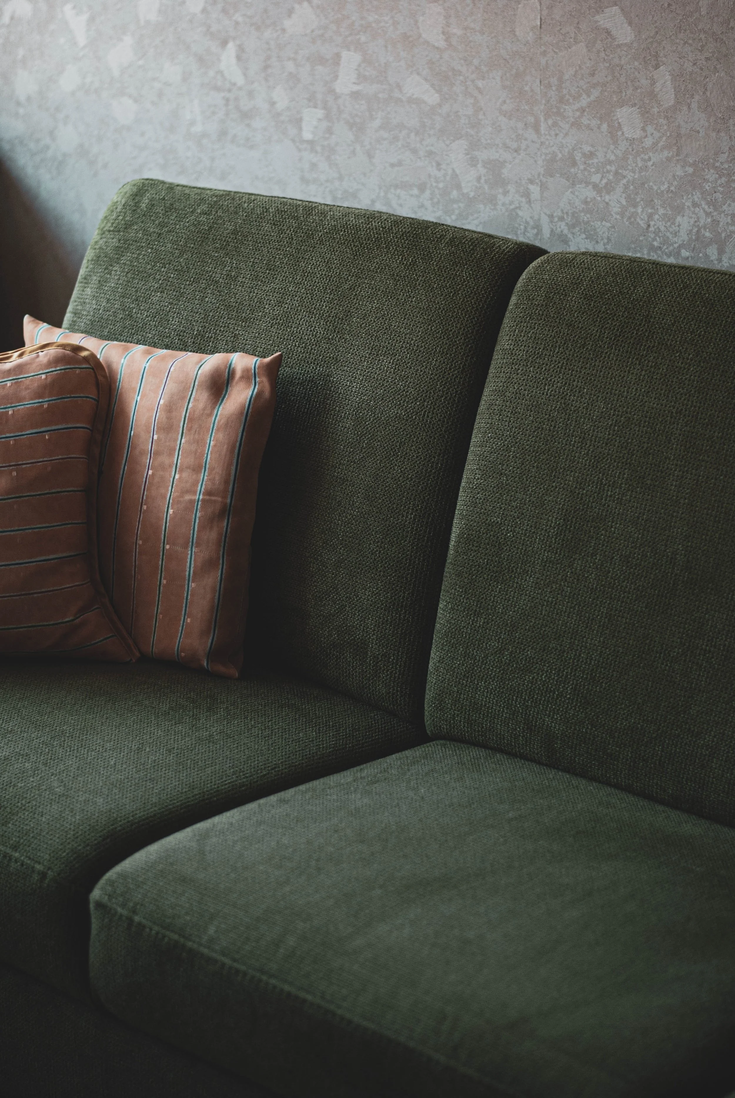 Close-up of a dark green upholstered sofa with two striped pillows, against a textured wall.