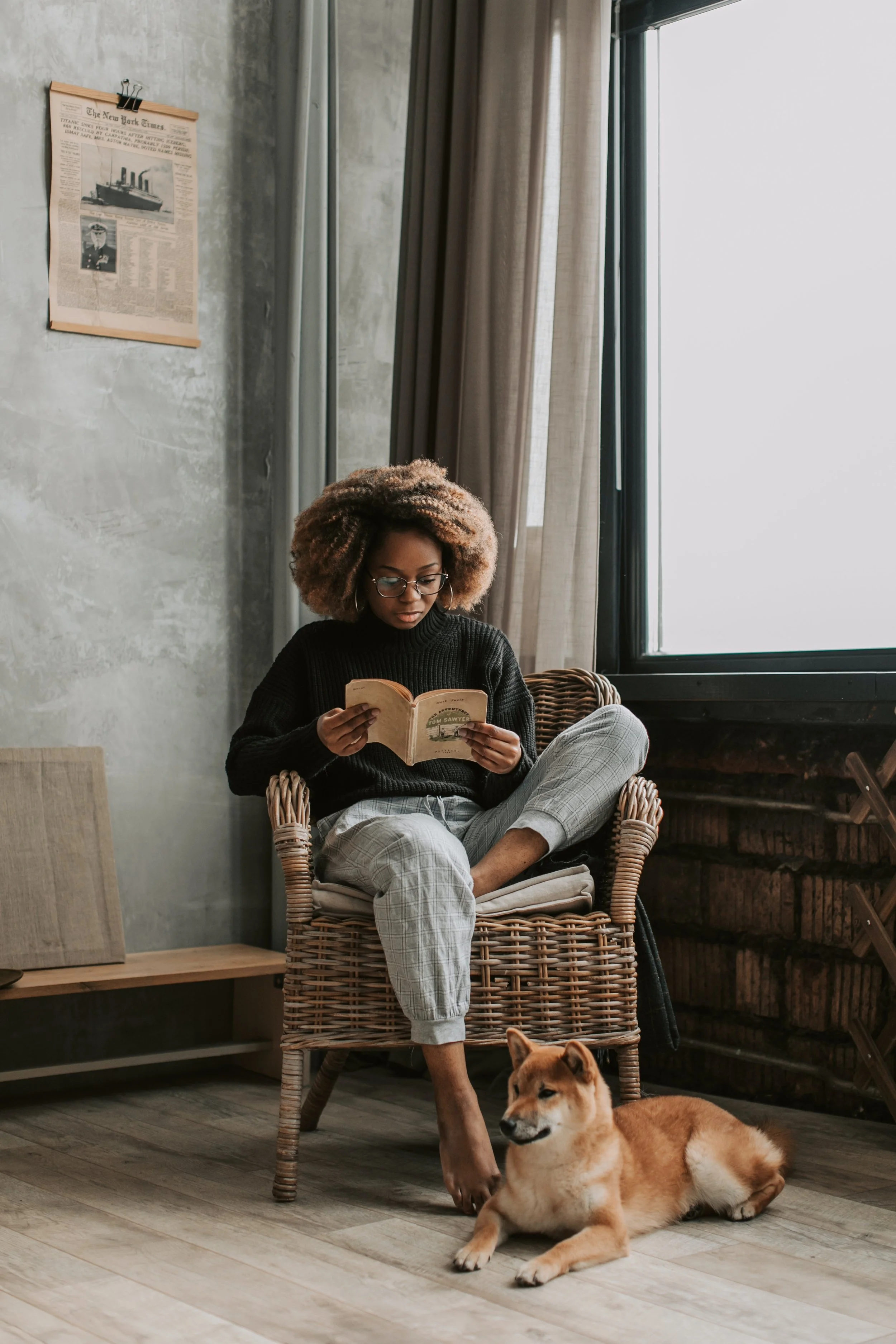 A woman with curly hair and glasses sitting in a wicker chair, reading a book, with a Shiba Inu dog lying beside her on the floor. The room has a large window, beige curtains, and a wall with a newspaper poster.