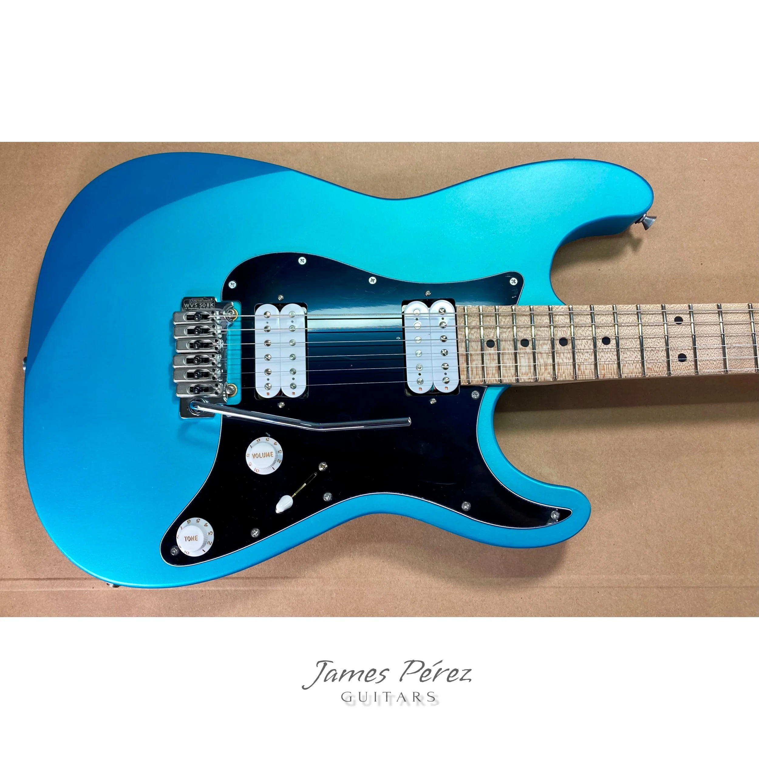 Blue electric guitar with black pickguard, two humbucker pickups, and a map of the fretboard laid on a brown surface, with the text "James Pérez GUITARS" below.