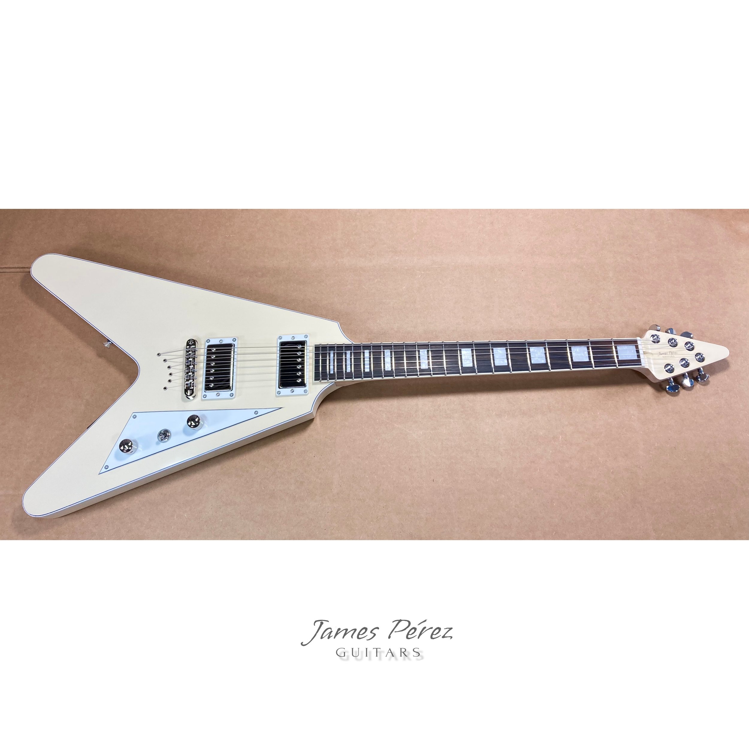 White electric guitar with a sharp, angular body shape and chrome hardware, placed on a brown surface, with the text "James Pérez GUITARS" at the bottom.